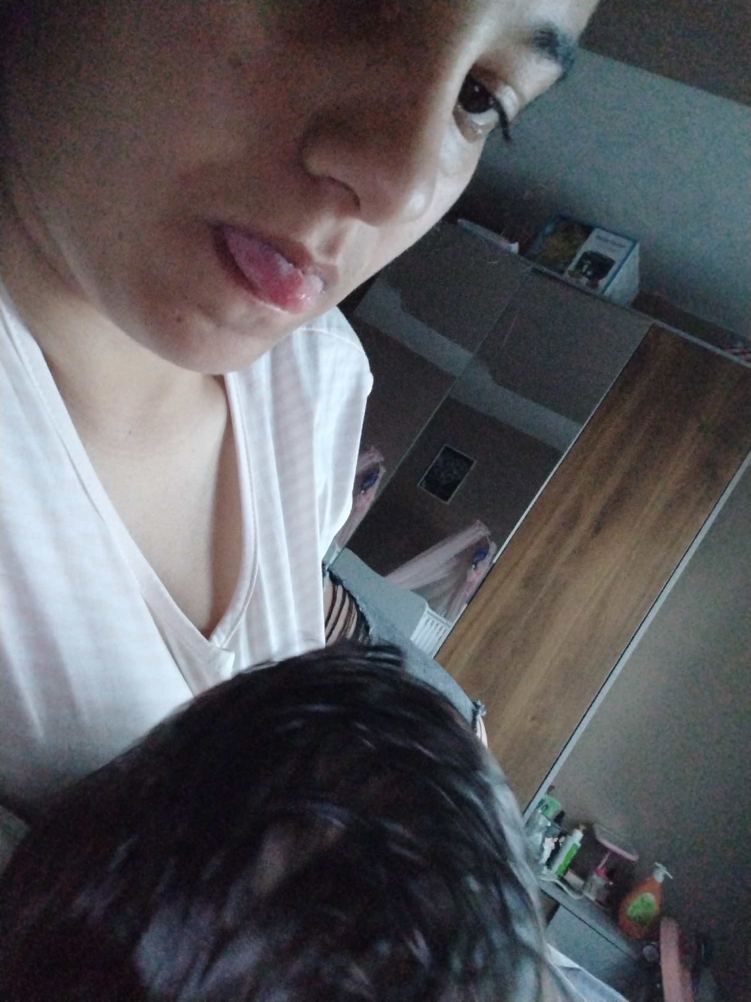 A close-up selfie depicts a person with dark hair, fair skin, and brown eyes. The individual is wearing a white V-neck shirt. The tongue is slightly protruding from the mouth. The background includes a mirrored wardrobe, revealing a reflection of what appears to be a crib or bed and other objects. Some cosmetic items can be seen on a surface beside the wardrobe. The location is unspecified.
