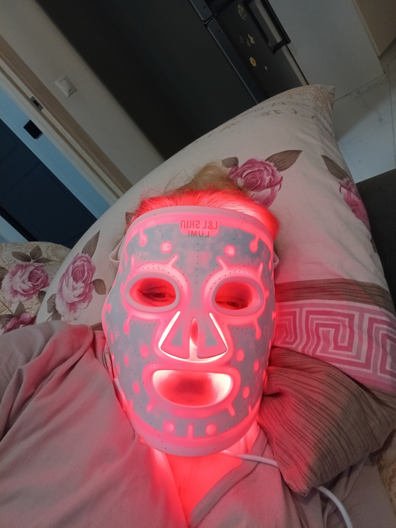 The image depicts an individual lying in an indoor residential setting, utilizing an LED light therapy facial mask. The subject is prone, with light-colored hair visible above the mask, and is wearing light-colored upper body clothing. The white facial mask, branded "L&L SKIN LUMI" (text inverted), emits a prominent red light, illuminating the subject's face, neck, and portions of the surrounding environment. A white cable extends from the lower right side of the mask. The subject rests on pillows; one features a pattern of pink roses and green leaves, while another exhibits a pink and white geometric design. In the background, a light-colored wall with a white electrical switch is visible. Dark-colored furniture or architectural elements, possibly including a cabinet with small magnets attached, are present in the deeper background. The scene is consistent with a private residence. Based on the visual evidence, a specific location within the city of Odintsovo, Russia, cannot be determined beyond an indoor dwelling.