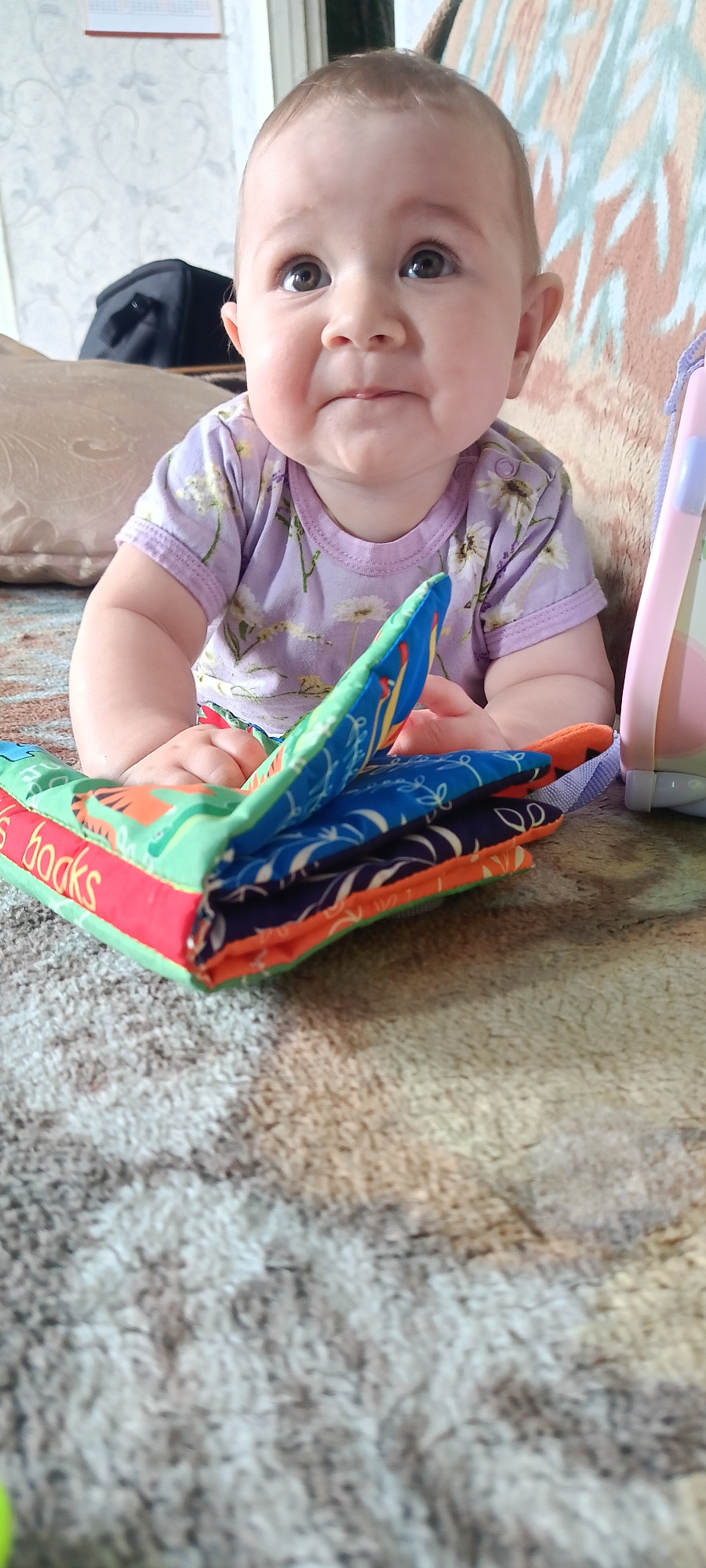 A baby sits on a patterned rug, facing forward with their head tilted upwards and to the left. The baby is wearing a light purple t-shirt with a floral pattern and has wide, dark eyes, a small nose, and slightly pursed lips. Their hands are resting on a folded fabric book with colorful patterns and red trim, on which the word "books" is partially visible in red lettering. Behind the baby, a light-colored couch with a textured pattern is visible. To the left of the baby, a black bag is partially visible. To the right, a pink and blue object, possibly a toy or a stroller part, is present. The scene appears to be indoors, in what could be a living room or play area, with a textured rug covering the floor and a patterned fabric in the background. The location is described as Каменное, Russia.