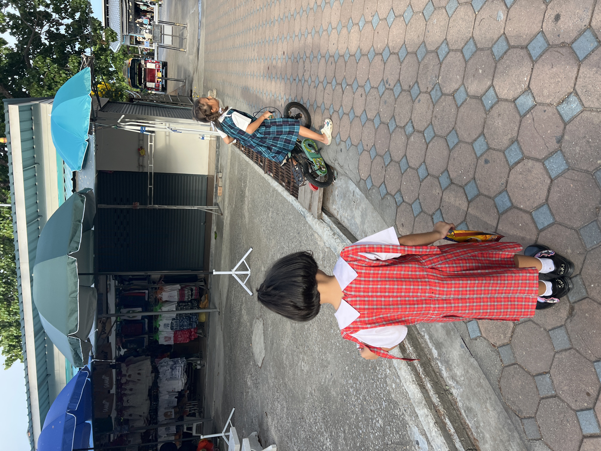 The image depicts a scene outside a row of shops in Baan Plai Laem, Thailand. Two young girls, dressed in school uniforms, are the primary subjects. In the foreground, a girl with dark hair stands facing away from the camera, wearing a red and white plaid pinafore over a white shirt. She is holding a yellow bag or package in her right hand. Further down the paved walkway, a second girl with braided hair rides a small green and black bicycle. She is wearing a blue and green plaid dress with a white shirt underneath and light-colored Crocs. She is looking to her right, away from the camera.
The environment consists of a concrete sidewalk with an octagonal and square tile pattern. Behind the girls, several market stalls are visible, covered by blue, green, and turquoise umbrellas. The stalls display clothing on racks. In the background, a red tuk-tuk vehicle is parked, and trees and other structures of the town can be seen. The lighting suggests it is daytime, possibly late afternoon, with shadows cast on the ground.