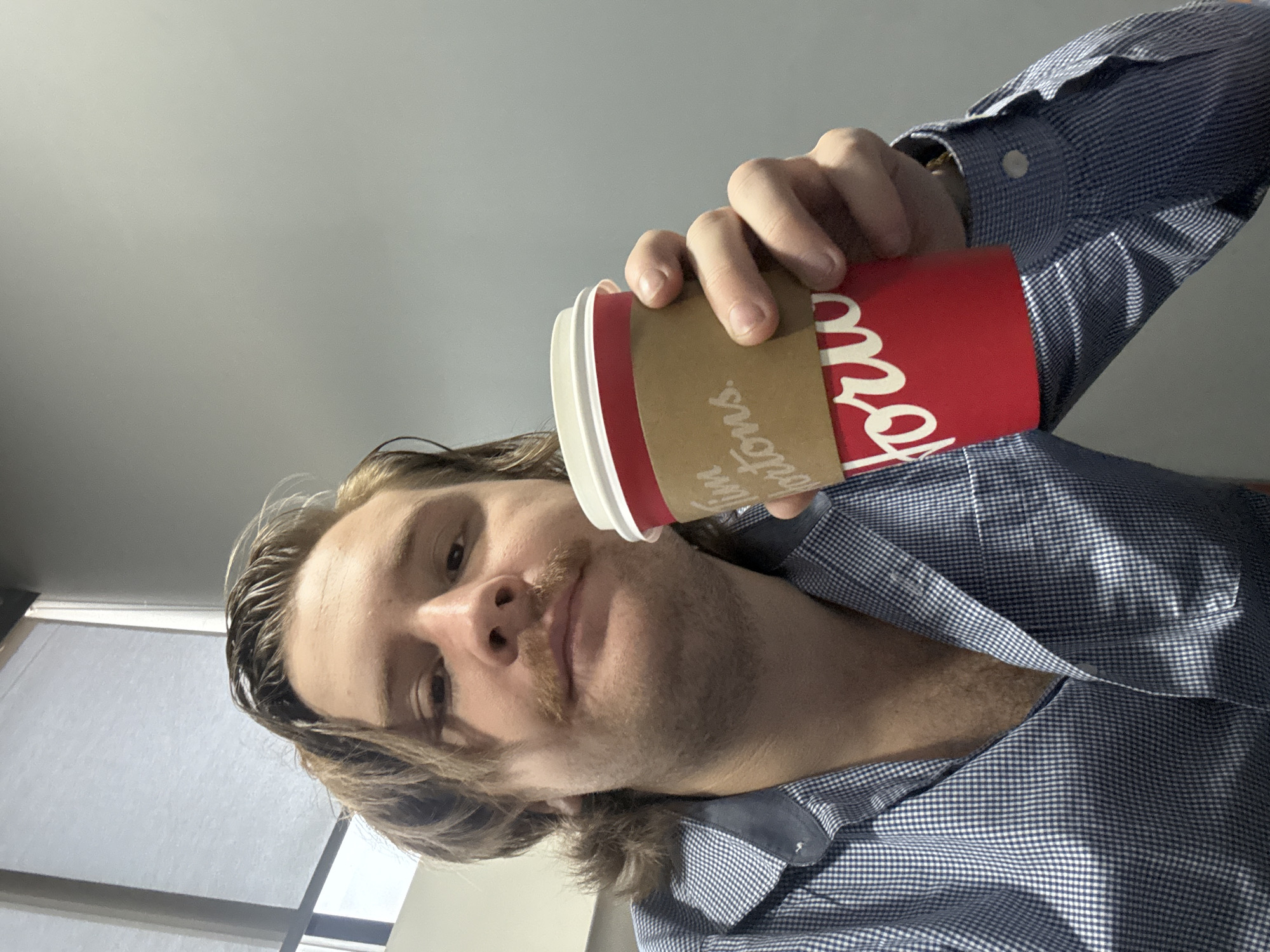 A low-angle selfie captures a man looking directly at the camera. He has medium-length, wavy brown hair that falls around his face and a prominent light brown mustache. In his right hand, raised towards the upper right of the frame, he holds a red and white disposable coffee cup. The cup features a brown cardboard sleeve with the distinctive "Tim Hortons" logo and the partial word "ations" visible in white lettering.
The man is wearing a blue and white checkered button-down shirt, unbuttoned at the top, revealing a glimpse of his chest. His expression is neutral, with a slight downward gaze towards the camera. The background consists of a plain, light-colored interior wall or ceiling, with a muted window visible on the far left, suggesting an indoor setting with ambient light. The overall image is tilted, emphasizing the upward angle from which it was taken, likely by the subject himself.