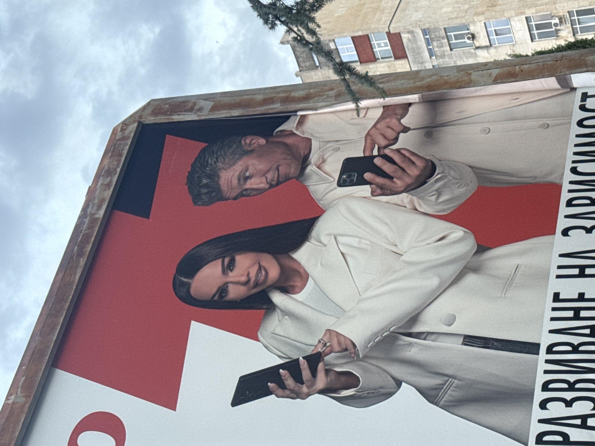 This image depicts a billboard advertisement featuring a man and a woman, both holding smartphones. The billboard has a red and white background with some Cyrillic text at the bottom that reads "РАЗВИВАНЕ НА ЗАВИСИМОСТ". The man, on the right, has short, styled gray hair and is wearing a light-colored collared shirt over a t-shirt. He is looking to his left, with a slight smile, and is holding a black smartphone with a carbon fiber case. The woman, on the left, has long, dark hair and is wearing a white blazer over a white top. She is also holding a black smartphone and looking forward with a smile, her left hand resting on the phone. The billboard is framed by a rusty metal structure. In the background, a multi-story building with several windows and air conditioning units is visible, along with some tree branches in the foreground. The sky is overcast with gray clouds. The billboard is located in Stara Zagora, Bulgaria, near the main pedestrian street (Glavna Ulitsa).