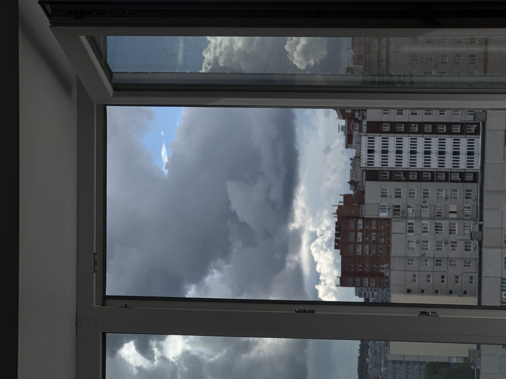 This media file captures a view from inside a room, looking out through a window at an urban landscape under a dramatic sky. The image is oriented vertically (rotated 90 degrees clockwise), showing the interior wall on the left, and the window frame in white or light grey.
The scene outside the window is dominated by a sky filled with heavy, dark grey clouds, suggesting stormy or overcast weather. However, there are also patches of clear blue sky and brighter, whiter clouds, indicating a dynamic weather pattern, possibly before or after a rain shower.
Below the cloudscape, several multi-story residential buildings are visible. The most prominent building, positioned in the mid-ground, features a block-like architectural style with sections of dark reddish-brown and lighter grey facades, adorned with numerous rectangular windows. Other, less distinct, buildings can be seen in the distance, contributing to a dense urban setting. The upper pane of the window reflects more of the cloudy sky.
There are no people visible in the image, nor is any specific activity taking place. The time of day appears to be daytime, but the heavy cloud cover gives the light a subdued quality. The architectural style of the buildings suggests a city in Eastern Europe or Russia. No visible text is present in the image.