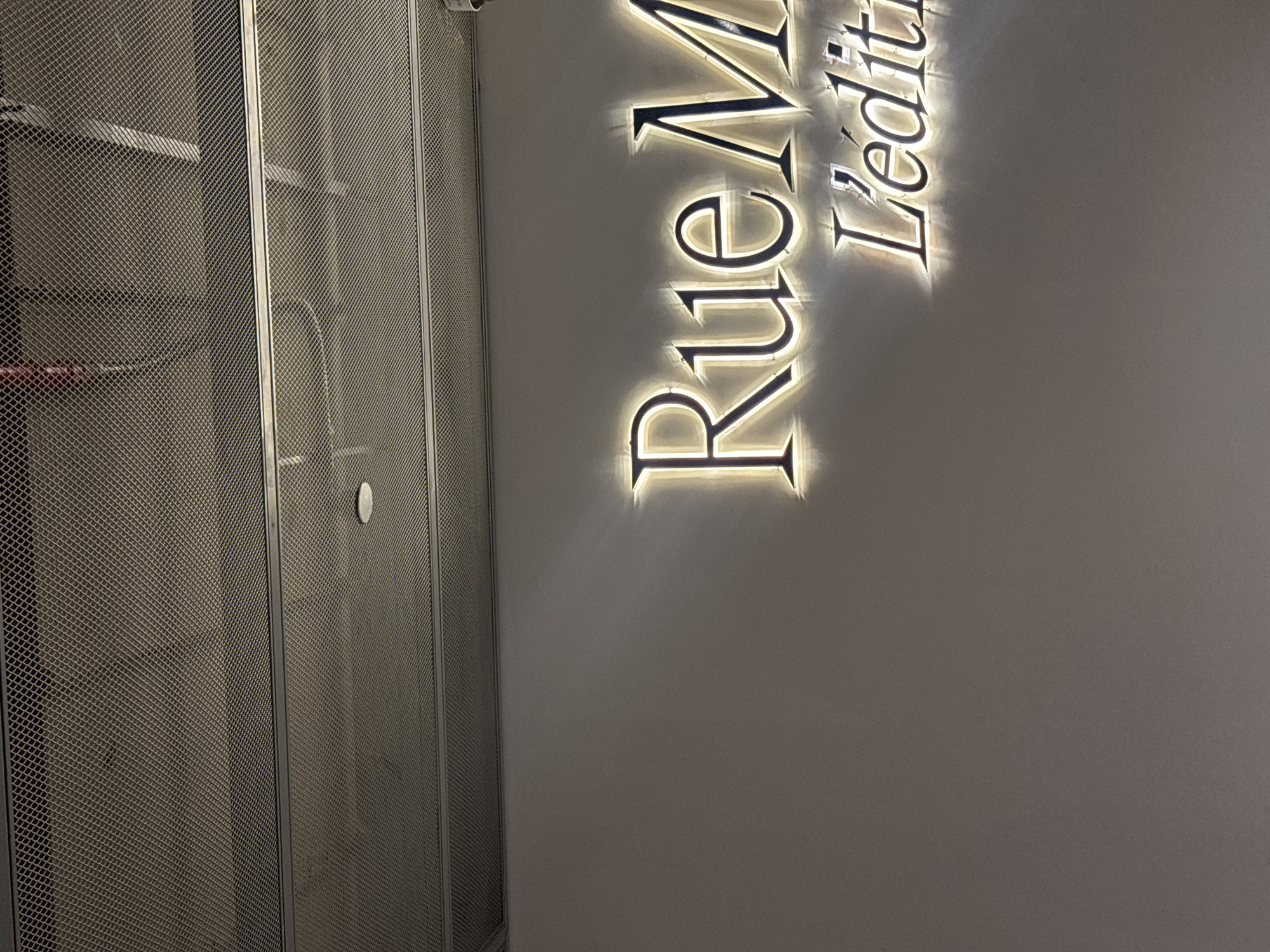 A dimly lit interior scene features a light-up sign on a neutral-colored wall. The sign displays two lines of text in a stylized, metallic font. The top line reads "Rue M" with illuminated edges, appearing in large, uppercase letters. Below it, in smaller, uppercase letters, is "L'edit", also with illuminated edges. Above the sign, a textured ceiling with a metallic grid pattern is visible, interrupted by horizontal beams and a round light fixture. The lighting from the sign casts a soft glow on the wall. The environment appears to be clean and modern. There are no people or animals visible in the image. The overall impression is that of a retail establishment or a place of business with a sophisticated aesthetic.