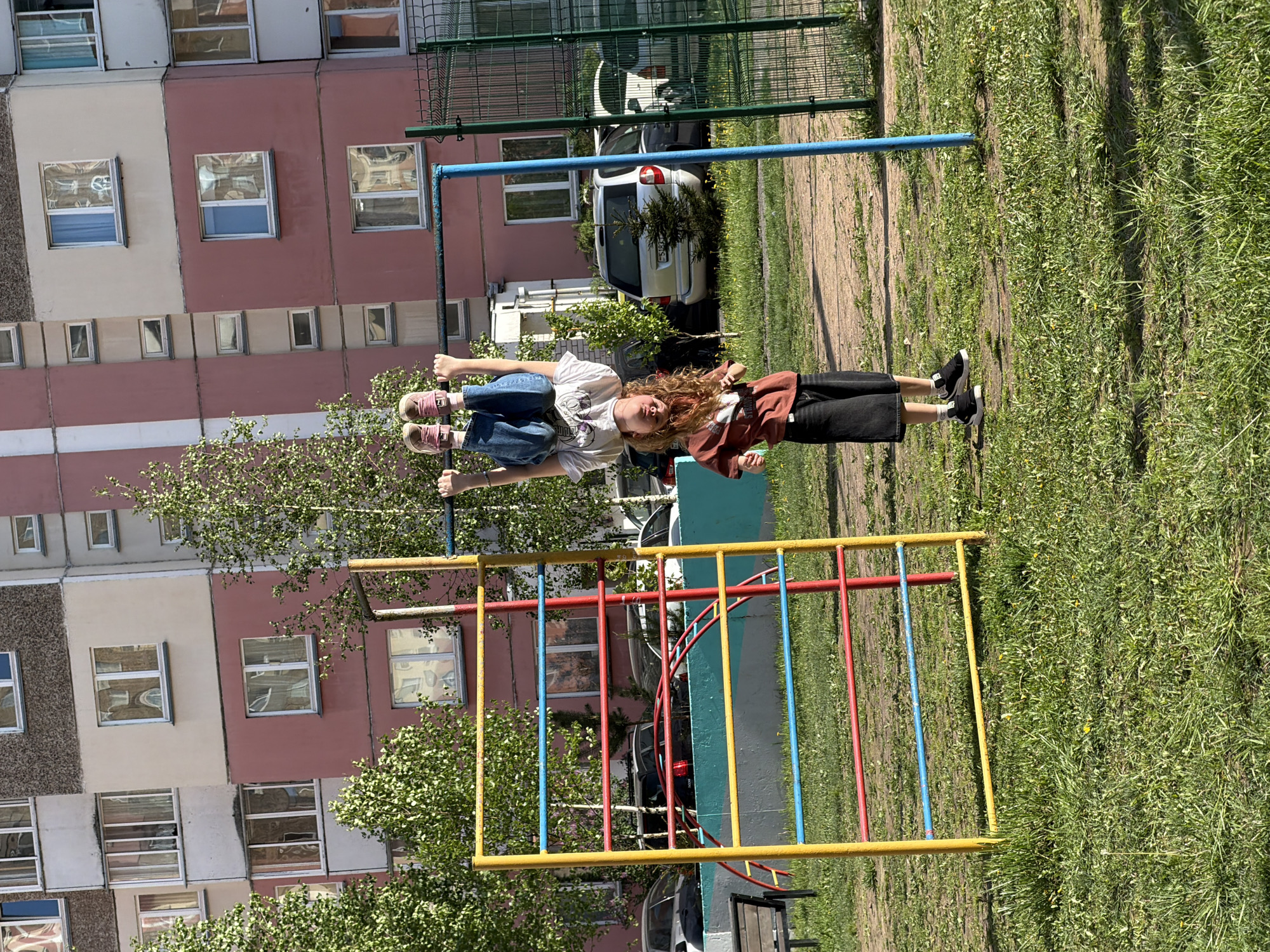 Two people are hanging upside down from monkey bars in an urban setting. A child with long, curly, reddish-brown hair is wearing a brown shirt, black shorts, and black shoes, hanging from one side. The other person, presumably a child, is wearing blue jeans, a white t-shirt, and pink shoes, and is hanging from the other side of the monkey bars. The monkey bars are multi-colored, with yellow, blue, and red bars. A building can be seen in the background, with multiple windows and a mix of beige, gray, and pink panels. There is grass and trees in the foreground and background. Cars are visible behind a fence in the background.