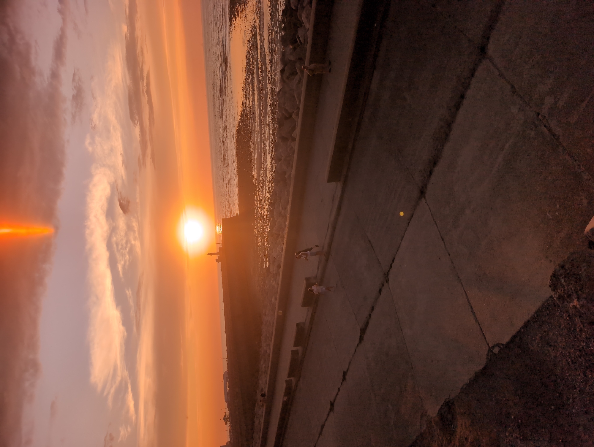 An eye-level, medium-shot captures a serene sunset over the coast of Le Tréport, France. The sky is a canvas of warm oranges and pinks, with streaks of gray clouds scattered across. A bright, glowing sun hangs low on the horizon, casting a shimmering reflection on the calm sea. In the distance, a lighthouse stands tall on the headland, a silent sentinel against the vibrant sky. The foreground is dominated by a concrete walkway, its weathered surface hinting at countless footsteps. A few figures are visible along the walkway: a small child in white runs towards the sea, while an adult, possibly their parent, walks beside them. Further along, another individual stands, seemingly observing the breathtaking view. The rocky shore, a mosaic of gray and brown, meets the water's edge, creating a natural barrier between the land and the sea.
