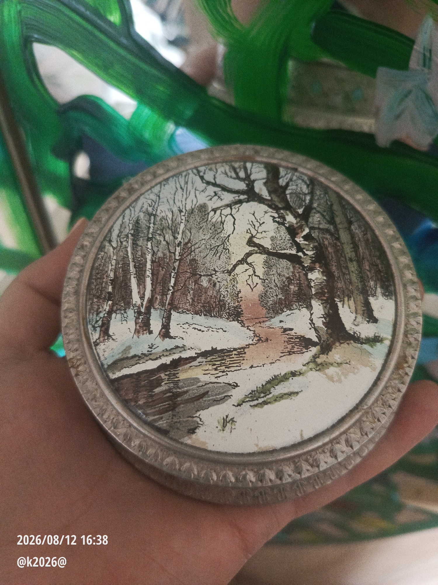 A circular, decorative object with a detailed illustration of a snowy forest scene is held in a person's hand. The illustration depicts bare trees with visible branches, patches of snow on the ground, and a path winding through the woods. The object has a metallic, patterned rim. The background of the image is blurred and appears to be an abstract painting with bold strokes of green. Text overlays in the bottom left corner read "2026/08/12 16:38" and "@k2026@". There is no discernible connection to the city of Minsk, Belarus, in the image content.