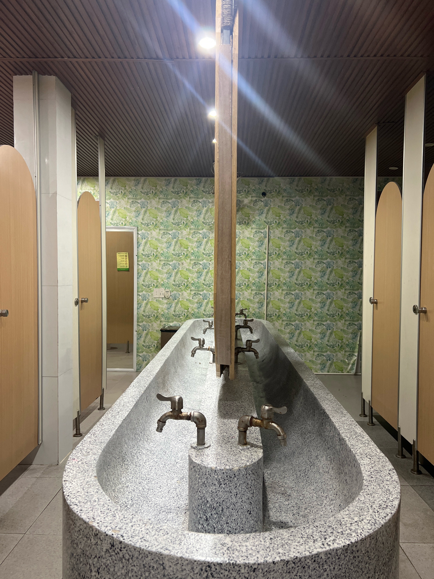 The image depicts a public washroom facility, likely in Phu Quoc, Vietnam. The primary focus is a long, trough-like sink made of speckled gray material, with several brass faucets protruding from its surface. The sink is positioned in the center of the frame, leading the eye towards the rear of the room.
Flanking the sink are partitions, likely for individual stalls or changing areas, constructed from light-colored wood with rounded tops. These partitions are separated by white pillars. The walls behind the sink are adorned with a vibrant green and white tropical leaf patterned wallpaper.
In the background, a door with a yellow sign is visible, suggesting an entrance to another part of the facility. The ceiling is constructed of dark, ribbed panels, with overhead lighting fixtures casting beams of light downwards. The floor is tiled with gray square tiles.
The overall impression is one of a functional, albeit somewhat dated, public amenity. The tropical wallpaper suggests a location that embraces its environment. The scene is static, with no people or ongoing actions visible, presenting a snapshot of the washroom's architecture and design.