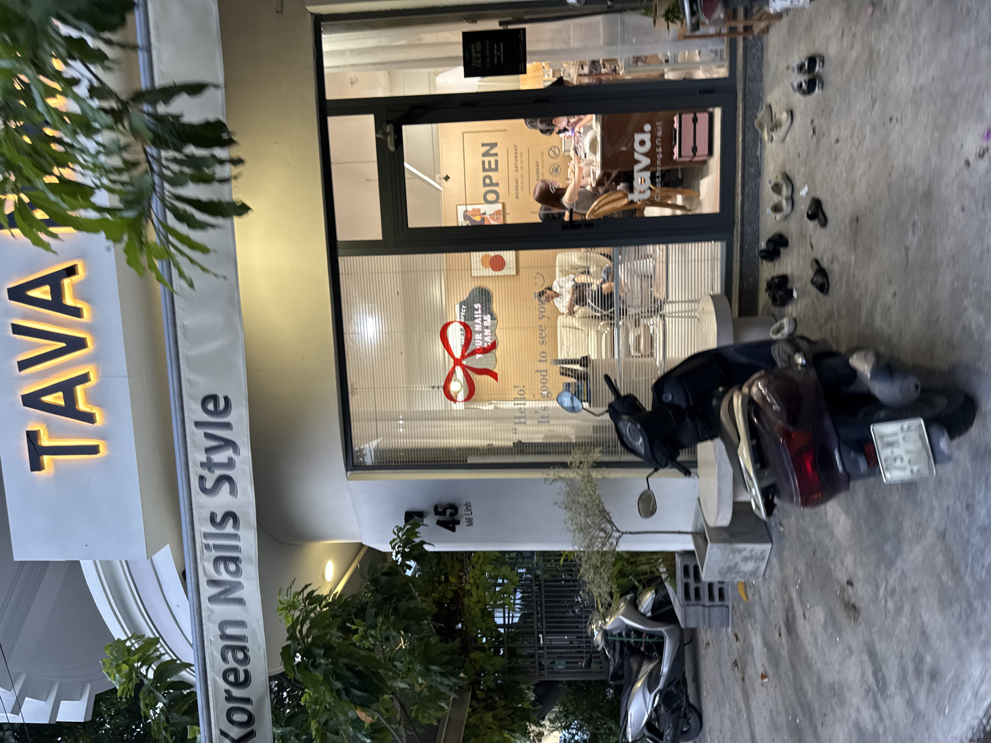 This image shows the exterior of a nail salon in Nha Trang, Vietnam, named "TAVA Korean Nails Style". The salon has a prominent illuminated sign reading "TAVA" in large orange letters. Below this, a smaller banner reads "Korean Nails Style". The salon entrance features a large glass door and window, revealing the interior where several people are engaged in nail services. A sign inside the window displays "OPEN". The street outside the salon is paved with concrete and several parked scooters are visible. A few pairs of shoes are placed on the ground in front of the salon entrance. The building appears to be a multi-story structure with a white facade. The address "45 Mê Linh" is displayed on the wall next to the entrance.