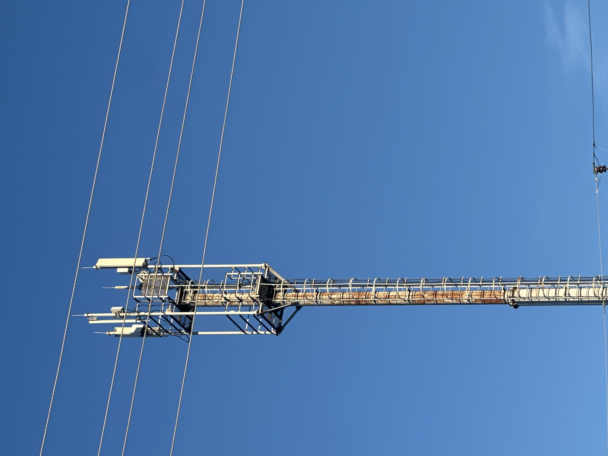 An outdoor, medium-shot, vertical, eye-level, slightly-low-angle, full-body photograph captures a cell tower against a clear, bright blue sky in Gavrilov Posad, Russia. The cell tower, a tall, slender, metal structure, is positioned off-center to the left of the frame. It is comprised of a white, lattice-like frame extending upwards. Several white, rectangular antennas are mounted on the upper section of the tower. A vertical ladder with white rungs runs along the length of the tower. The tower's central pole exhibits significant rust and discoloration, indicating age or exposure to the elements. Four parallel, thin, metallic wires run horizontally across the upper portion of the frame, passing in front of the cell tower and the sky. A portion of another wire structure with small, dark electrical components is visible in the lower right corner of the image. The scene is devoid of any people or animals.