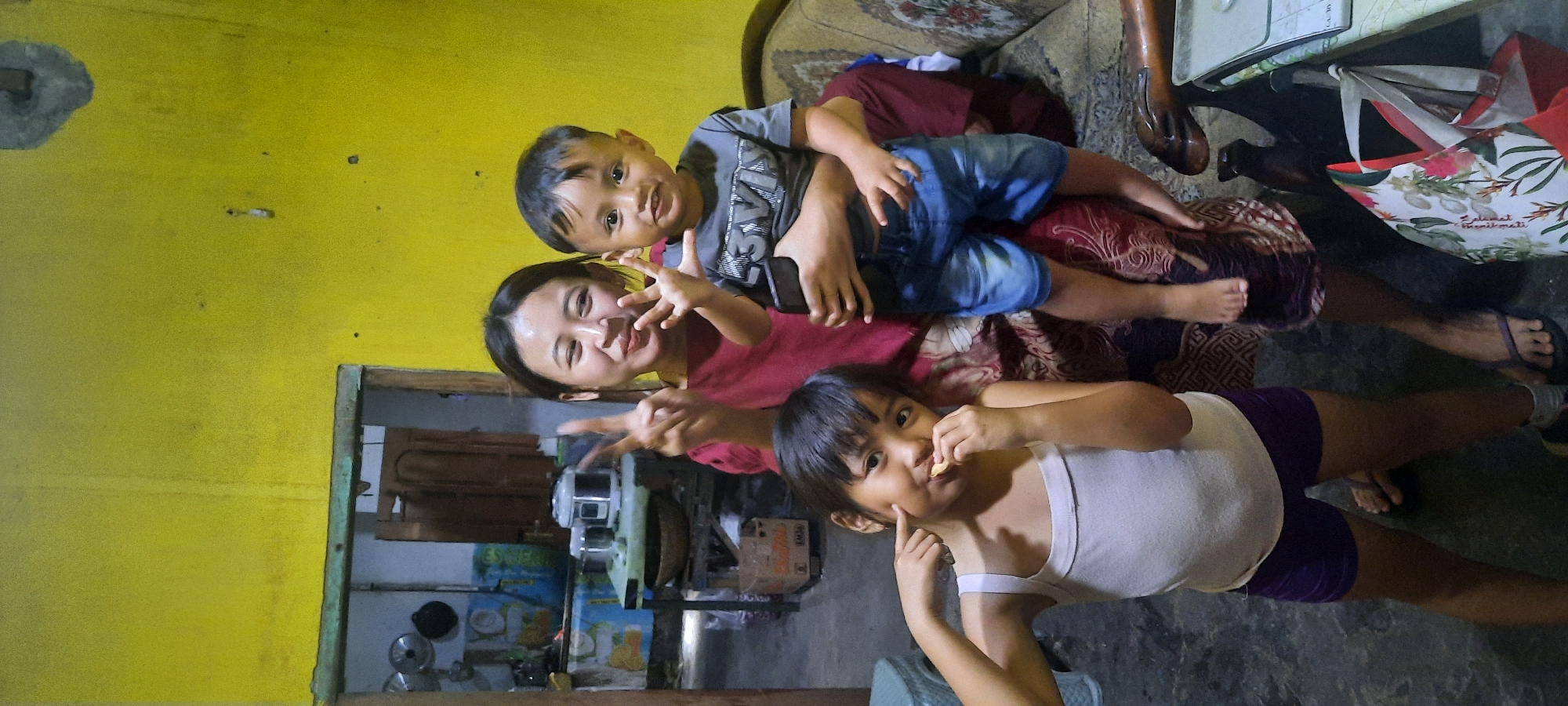This image shows three people, two children and one adult, in what appears to be a home environment in Selopuro, Indonesia. A young woman, likely the mother, is in the foreground, looking at the camera with a playful expression, making a peace sign with her fingers. She is wearing a red shirt. A young boy is being held by the woman, sitting on her lap or shoulders, and he is also looking towards the camera, smiling. He is wearing a grey t-shirt and blue denim shorts. Another young child, a girl, is positioned lower in the frame, looking towards the camera and touching her cheek. She is wearing a white tank top and purple shorts. The scene is indoors, with a yellow wall visible in the background. There are hints of furniture, including a couch and what looks like a table with some items on it, such as a patterned bag with floral designs. The flooring appears to be concrete or tile. The overall mood is casual and happy, suggesting a family moment captured for a photograph. There is no visible text in the image.