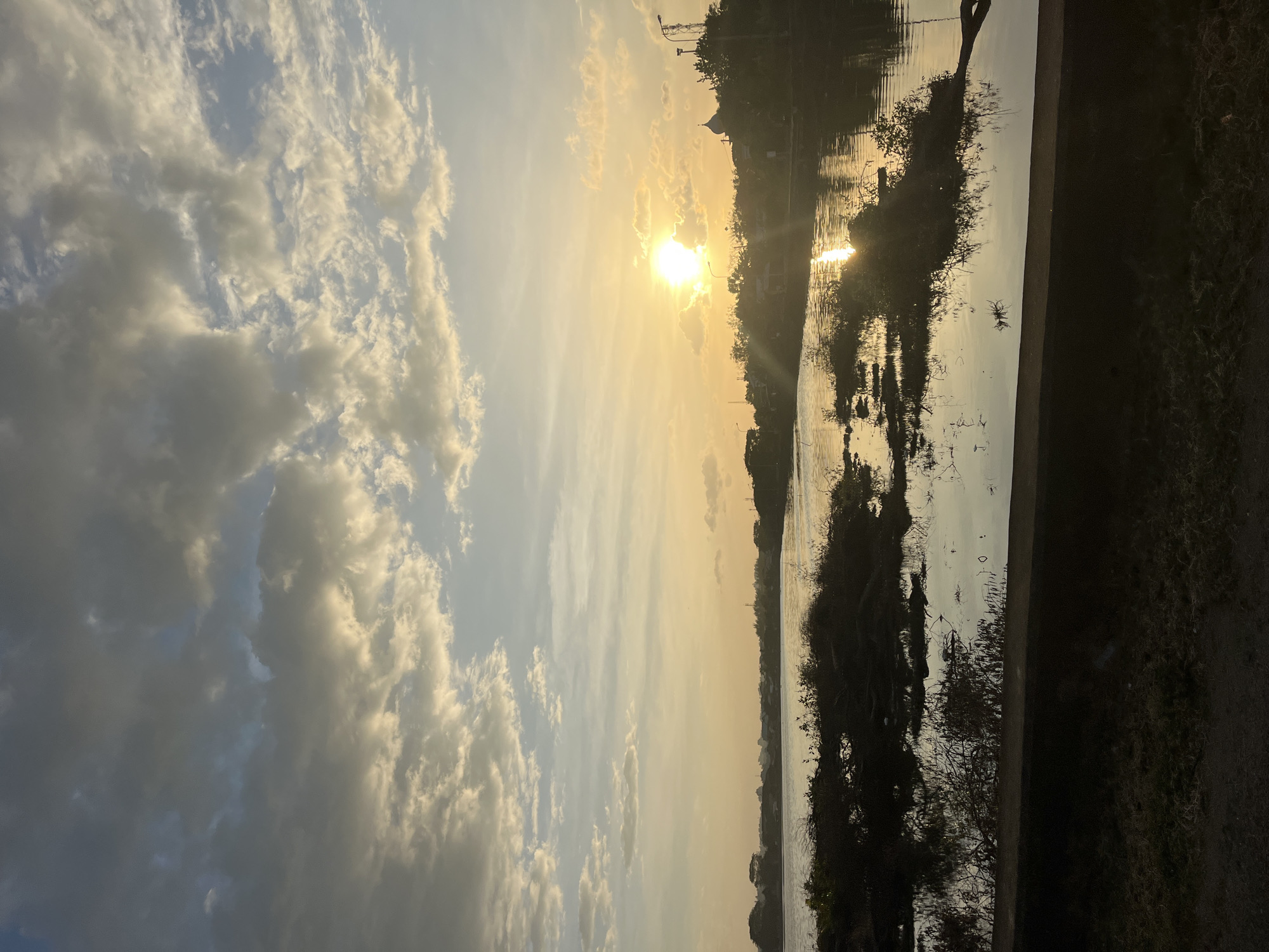 The image shows a sunset over a body of water. The sun is a bright spot in the sky, casting a golden light across the scene. The sky is filled with clouds, some white and fluffy, others darker and more ominous. The water reflects the sky, creating a mirror image of the clouds and the sun. There are trees and other vegetation along the shoreline. The image may have been taken in the city of Lhokseumawe, Indonesia, but there is no clear evidence to confirm this.