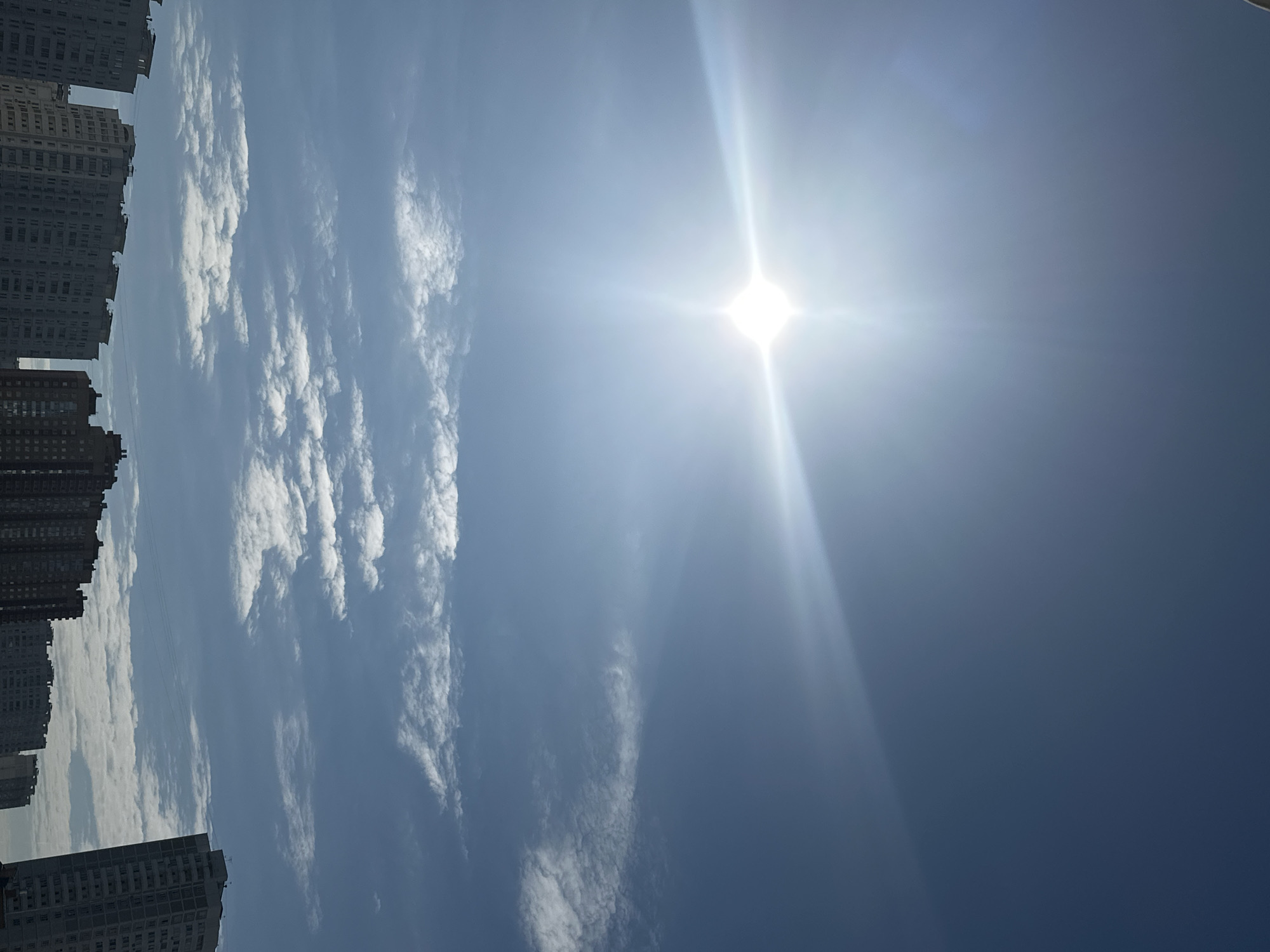 The image shows a clear blue sky with a bright sun, creating lens flare. There are scattered white clouds. In the foreground, several multi-story residential buildings are visible, with a skyline that suggests an urban or suburban environment. The scene is set in the city of Podolsk, Russia. There are no people or animals visible in the image. The architecture of the buildings indicates a typical Eastern European style of apartment blocks.