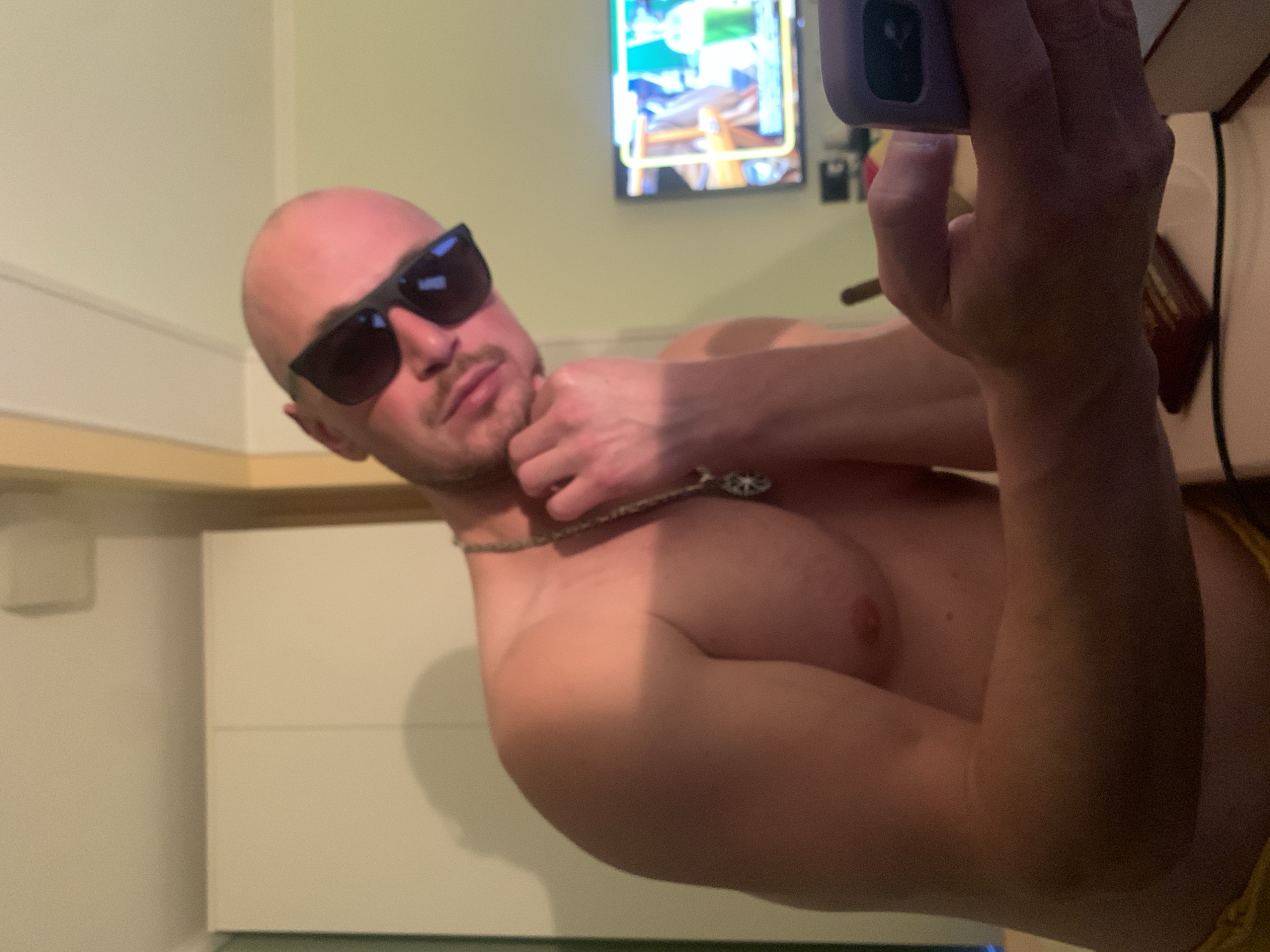 The media file shows a close-up, waist-up shot of a shirtless, bald man wearing black sunglasses and a necklace. He is holding a phone in his right hand, which is angled towards the camera as if taking a selfie. The man is positioned in what appears to be a domestic setting, possibly a bathroom or kitchen, with neutral-colored cabinets and a wall visible in the background. A television screen displaying an image with bright colors is mounted on the wall behind him. The lighting is dim, suggesting it may be evening or indoors with artificial light. The man's expression is partially obscured by his sunglasses, but he appears relaxed. The image is oriented vertically, rotated 90 degrees counter-clockwise from the typical portrait orientation. No text is visible in the image. The provided location context is Pushkin, Russia.