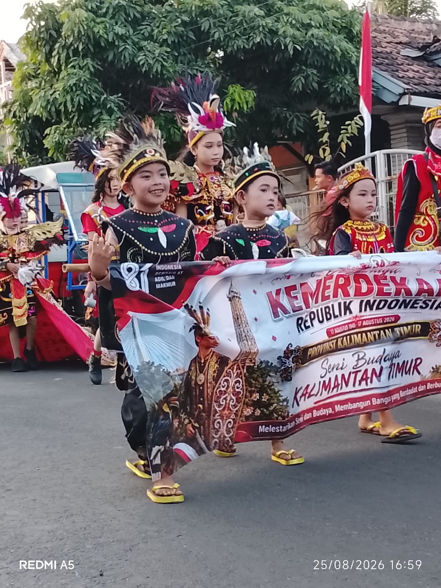 In Semanding, Indonesia, a group of children in traditional costumes participates in a parade. The children, some wearing elaborate headdresses and face paint, hold a banner that reads "KEMERDEKAN REPUBLIK INDONESIA." The banner also features text in Indonesian, including "PROVINSI KALIMANTAN TIMUR" and "Seni Budaya Kalimantan Timur." The children are walking on a paved road, with trees and buildings visible in the background. A red and white Indonesian flag is displayed on a pole to the right of the frame. The scene appears to be a celebration of Indonesian Independence Day.