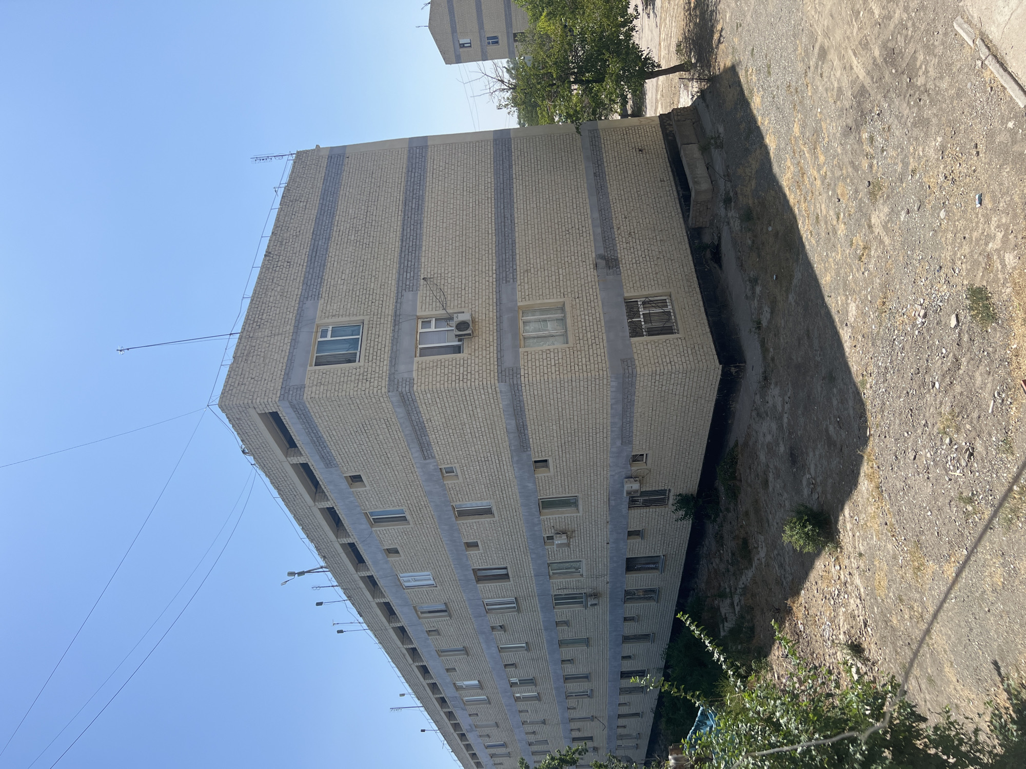 This image captures a view of a large, multi-story apartment building constructed with light-colored bricks and horizontal grey bands. The building has a flat roof with several antennas and a railing. The windows on the building are rectangular, and some have air conditioning units mounted outside. In the foreground, there is a dry, dusty ground with sparse vegetation and rocks. The background shows a clear blue sky and the upper portion of another similar apartment building and some trees. The scene appears to be in an urban or semi-urban environment, likely residential. The location is identified as Şamaldı-Say, Kyrgyzstan.