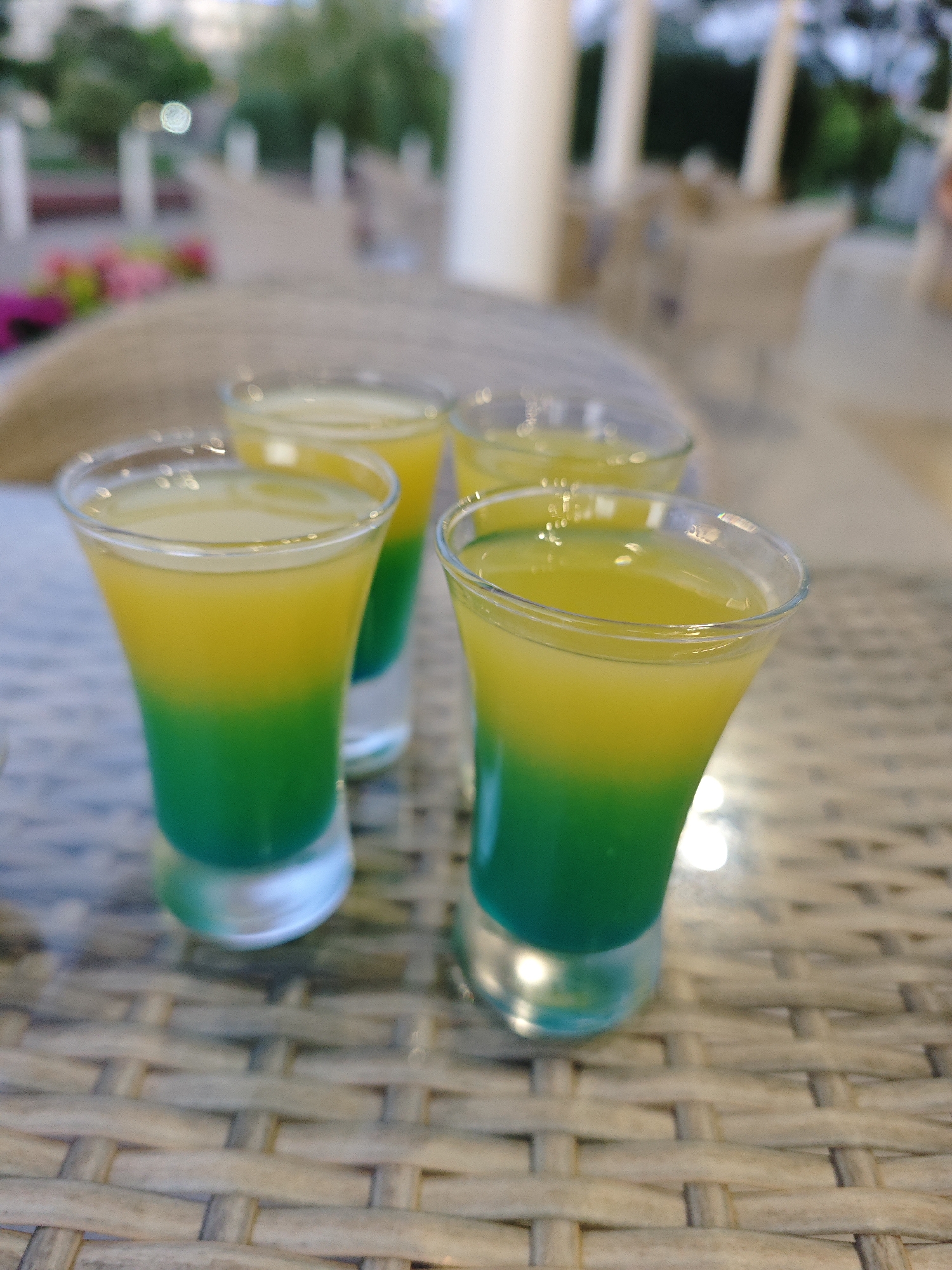 Four shot glasses containing layered green and yellow beverages are arranged on a wicker table. The shot glasses are tall and narrow, with the green liquid occupying the bottom half and the yellow liquid filling the top half. The background is blurred but suggests an outdoor setting with greenery, possibly a resort or cafe. The lighting is soft, with reflections visible in the glasses. The image does not provide enough information to determine the specific location in Sochi, Russia.
