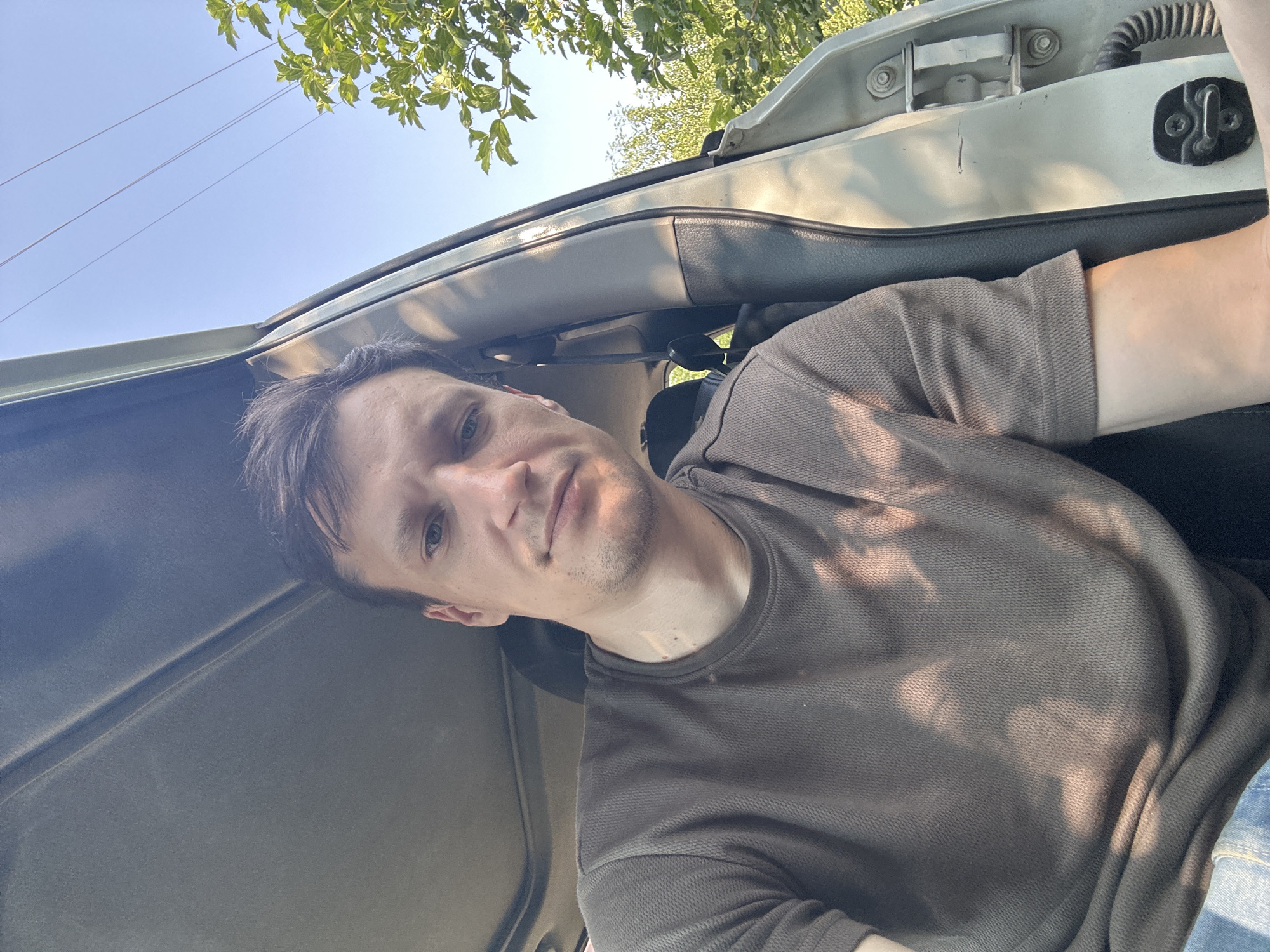 A man is sitting in the driver's seat of a car with the door open, looking at the camera. He has short brown hair and is wearing a dark brown t-shirt. The car's sunroof is visible, and sunlight is casting shadows on the man and the interior of the car. Outside the car, green trees and a blue sky are visible. The image was taken outdoors in Shatava, Ukraine.
