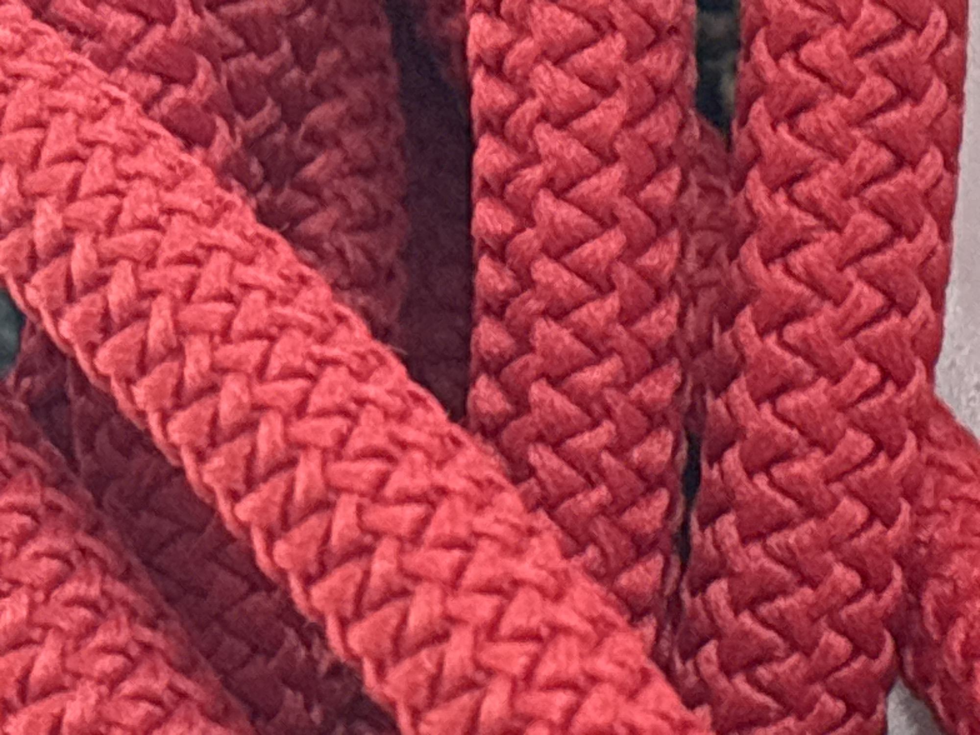 This is a close-up, macro photograph of braided red rope. The texture of the rope is highly detailed, showing the interwoven strands and the slight fuzziness of the material. The lighting creates subtle shadows that accentuate the three-dimensional weave. There are at least three distinct ropes or segments of rope visible, stacked diagonally across the frame. The background is dark and out of focus, providing no discernible details about the environment. There are no people, animals, or any other objects present in the image. The image does not provide any information about the location in the city of Zvenigorod, Russia, or any context outside of the rope itself.