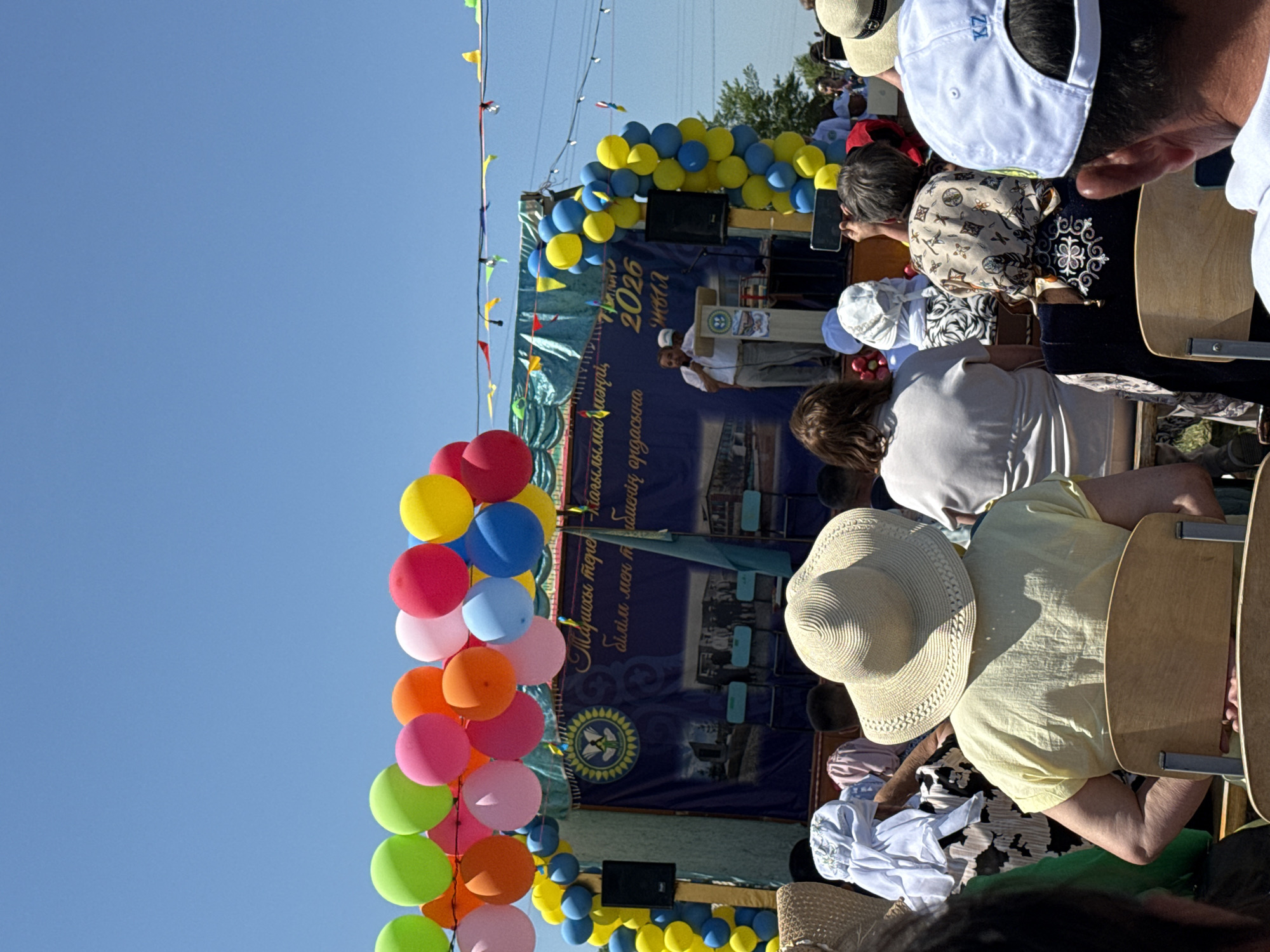 This image depicts an outdoor event in the city of Өндіріс, Kazakhstan, under a clear blue sky. The scene features a stage adorned with a banner, colorful balloons, and flags. In the foreground, a crowd of people, predominantly women, are seated and facing the stage. Many are wearing traditional attire, including headscarves and patterned garments. One woman in the foreground is wearing a wide-brimmed straw hat and a yellow top. On the stage, a man in a white shirt and pants is speaking at a podium, with another man standing behind him. The banner behind them contains text in Cyrillic script and an emblem with a circular design. The environment appears festive, with balloons and decorations suggesting a celebration or public gathering.