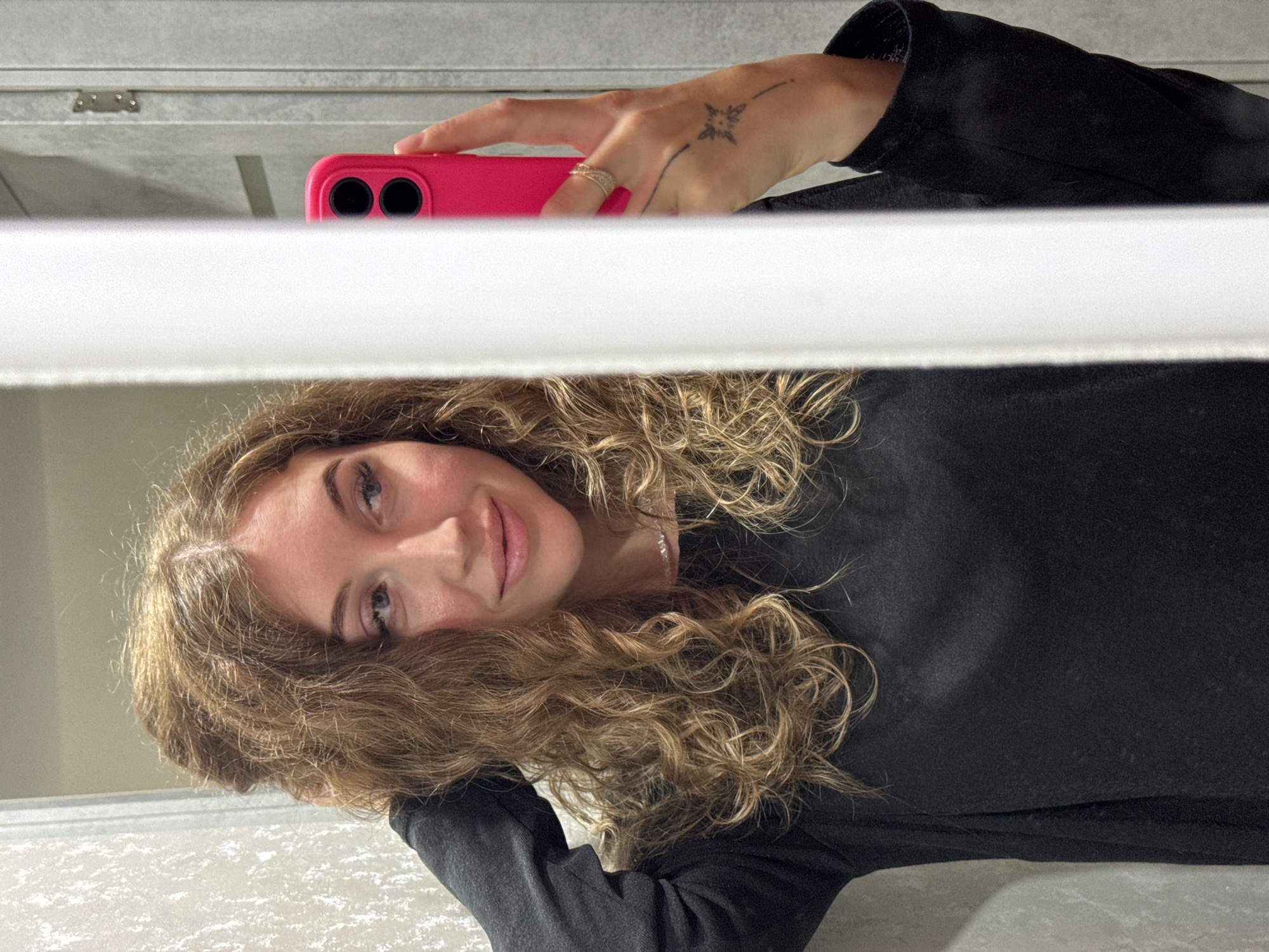 A selfie photograph shows a woman with curly brown hair, wearing a black long-sleeved shirt and holding a pink smartphone. Her right hand, adorned with a ring and a tattoo, is holding the phone. The background includes what appears to be a white blind and a gray wall. The location is not discernible from the image.