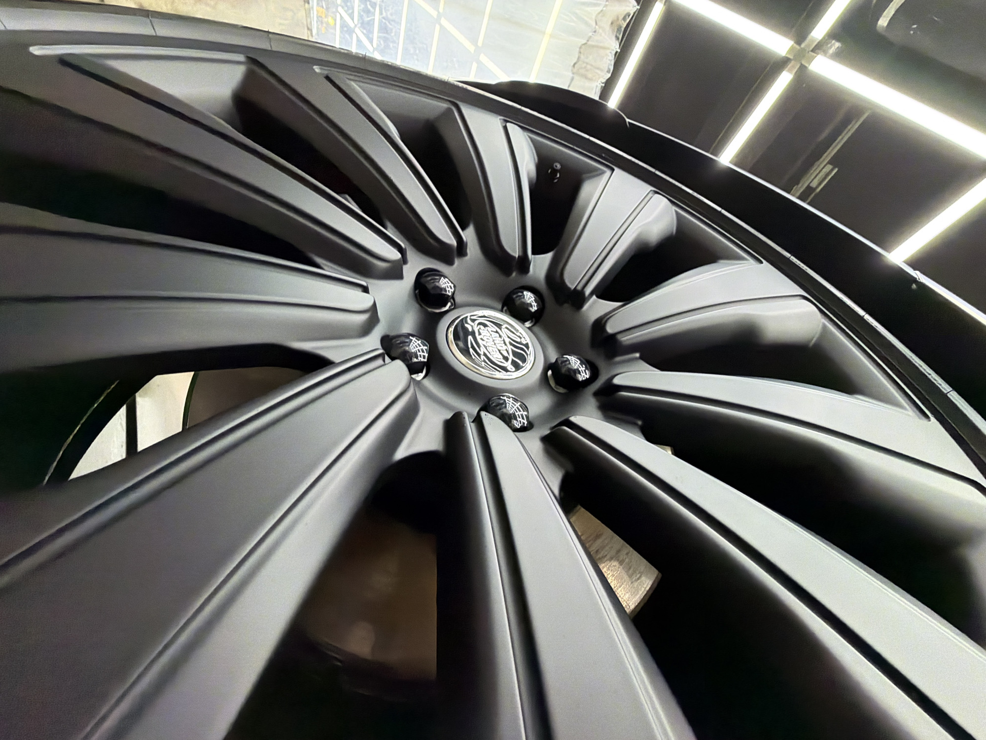 The image captures a detailed, low-angle close-up of a matte dark grey or black car wheel rim. The wheel features a striking multi-spoke design, with numerous robust spokes radiating outwards from the central hub. The finish appears uniform and immaculate, suggesting a recent cleaning, detailing, or application of a protective coating.
At the center of the wheel is a circular cap bearing a distinct logo. The emblem, visible in the detailed crops, clearly displays the "Land Rover" brand insignia with intricate silver-toned lettering and a crest-like design against a dark background. Five glossy lug nuts, possibly with a textured or patterned surface, surround the central cap, reflecting ambient light.
The setting is an indoor, professional environment, likely a garage, auto detailing studio, or showroom located in Krasnoyarsk, Russia. The background reveals a clean ceiling equipped with bright, linear fluorescent or LED light fixtures arranged in a grid pattern, which are prominently reflected on the glossy surfaces of the lug nuts and subtly on the upper portions of the wheel and the unseen car bodywork. No people are visible in the frame. The overall presentation emphasizes the pristine condition and aesthetic appeal of the wheel, indicating a focus on quality and detailed workmanship.