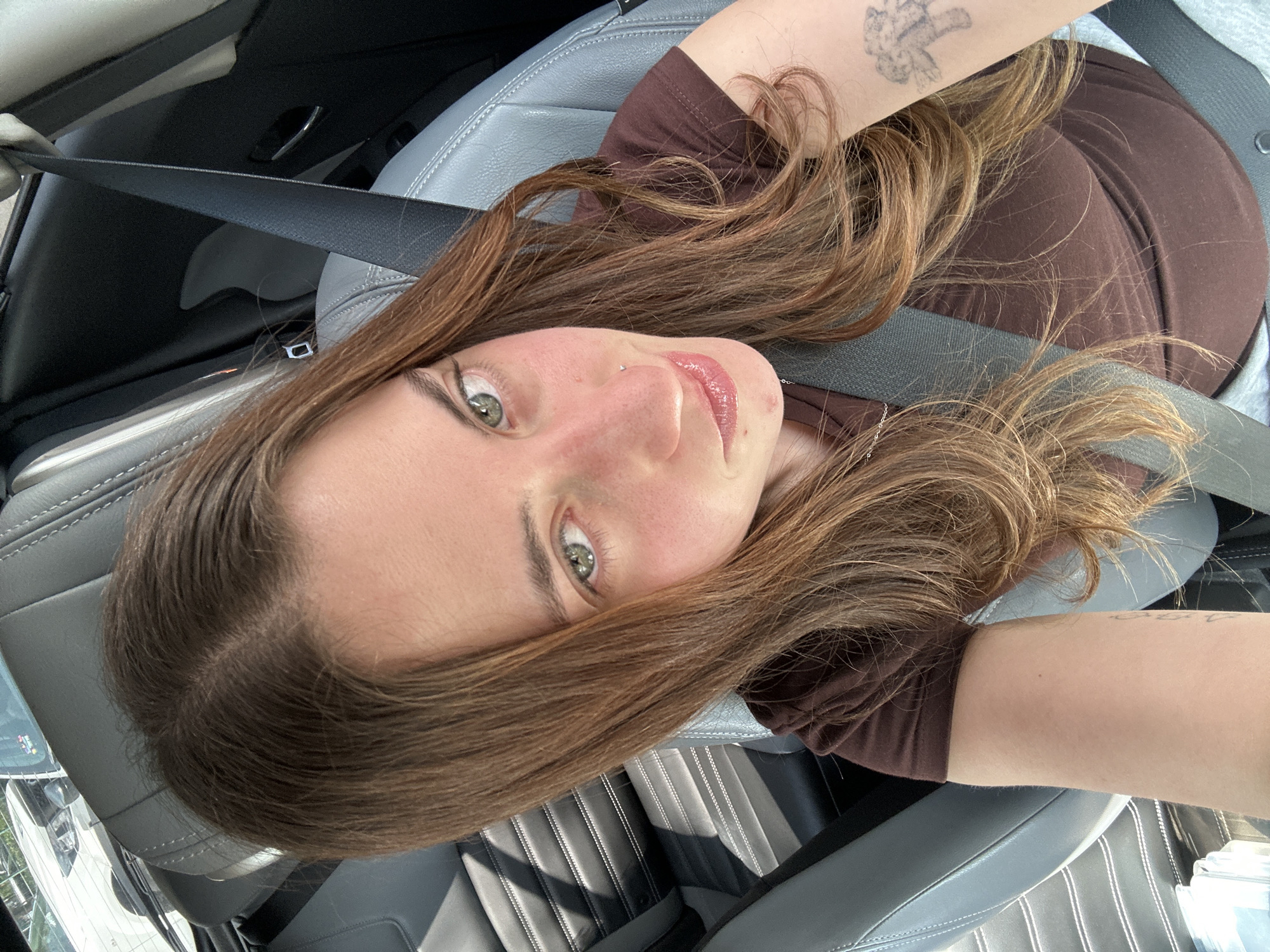 A woman is sitting in a car, wearing a brown shirt and a seatbelt. The interior of the car features gray leather seats with black accents. The location is likely inside a vehicle within the city of Huixquilucan de Degollado, Mexico. The woman is looking at the camera. She has a piercing on her nose. She also has a tattoo on her arm.