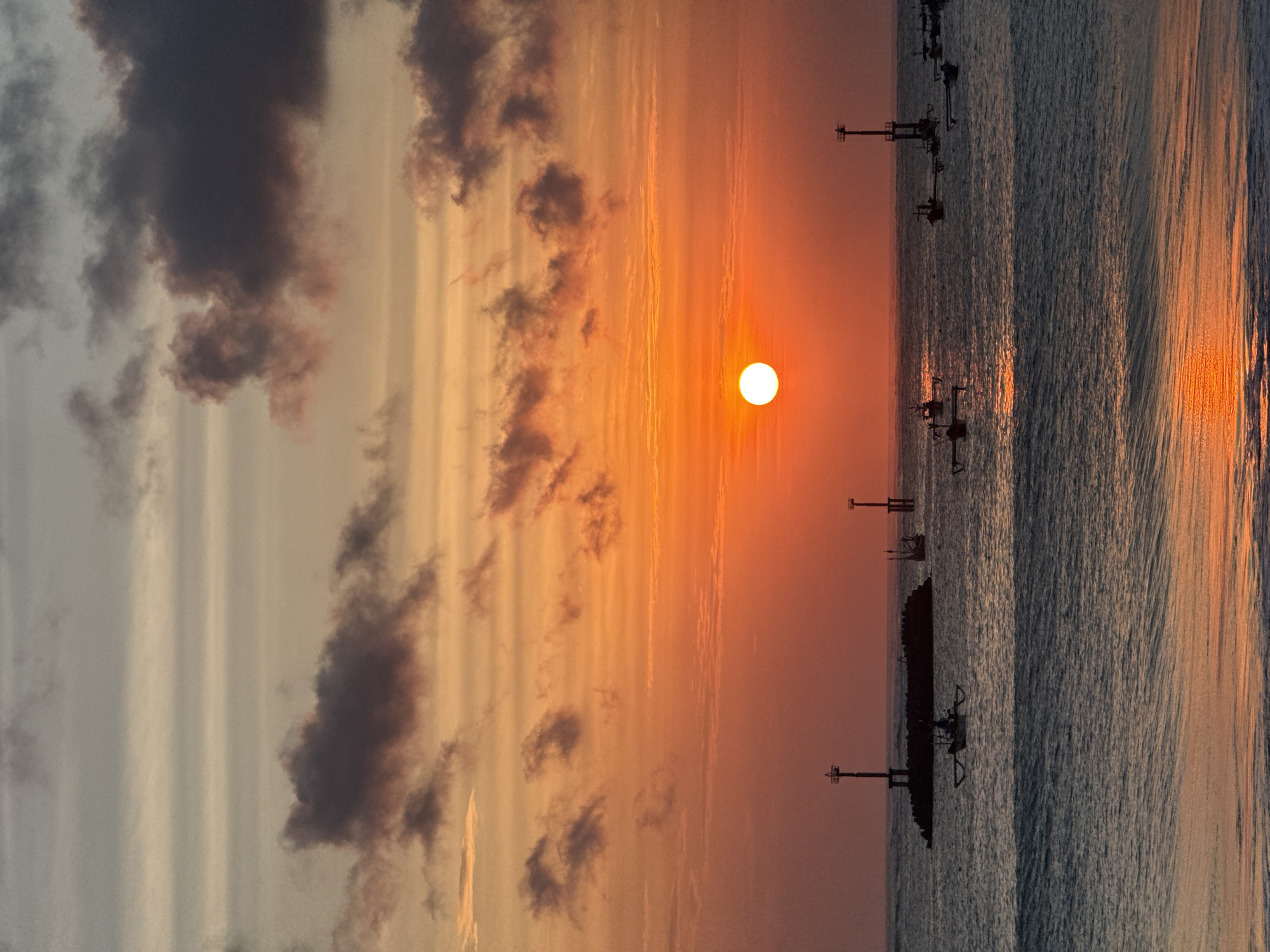 The image depicts a serene sunset over the ocean in Kuta, Indonesia. The sun, a bright orange orb, is low on the horizon, casting a warm glow across the sky, which is painted with hues of orange, yellow, and soft gray. Darker clouds are scattered across the upper portion of the sky.
In the middle ground of the ocean, several traditional Balinese fishing boats, known as "jukung," are visible. These boats, characterized by their outriggers, are scattered across the water. Two lighthouses stand tall, one prominently on a rocky breakwater, and another further out in the sea. The water itself is calm, with gentle waves rolling towards the shore. The reflection of the sunset can be seen shimmering on the water's surface. The overall atmosphere is tranquil and picturesque, capturing the essence of a coastal sunset in Kuta.