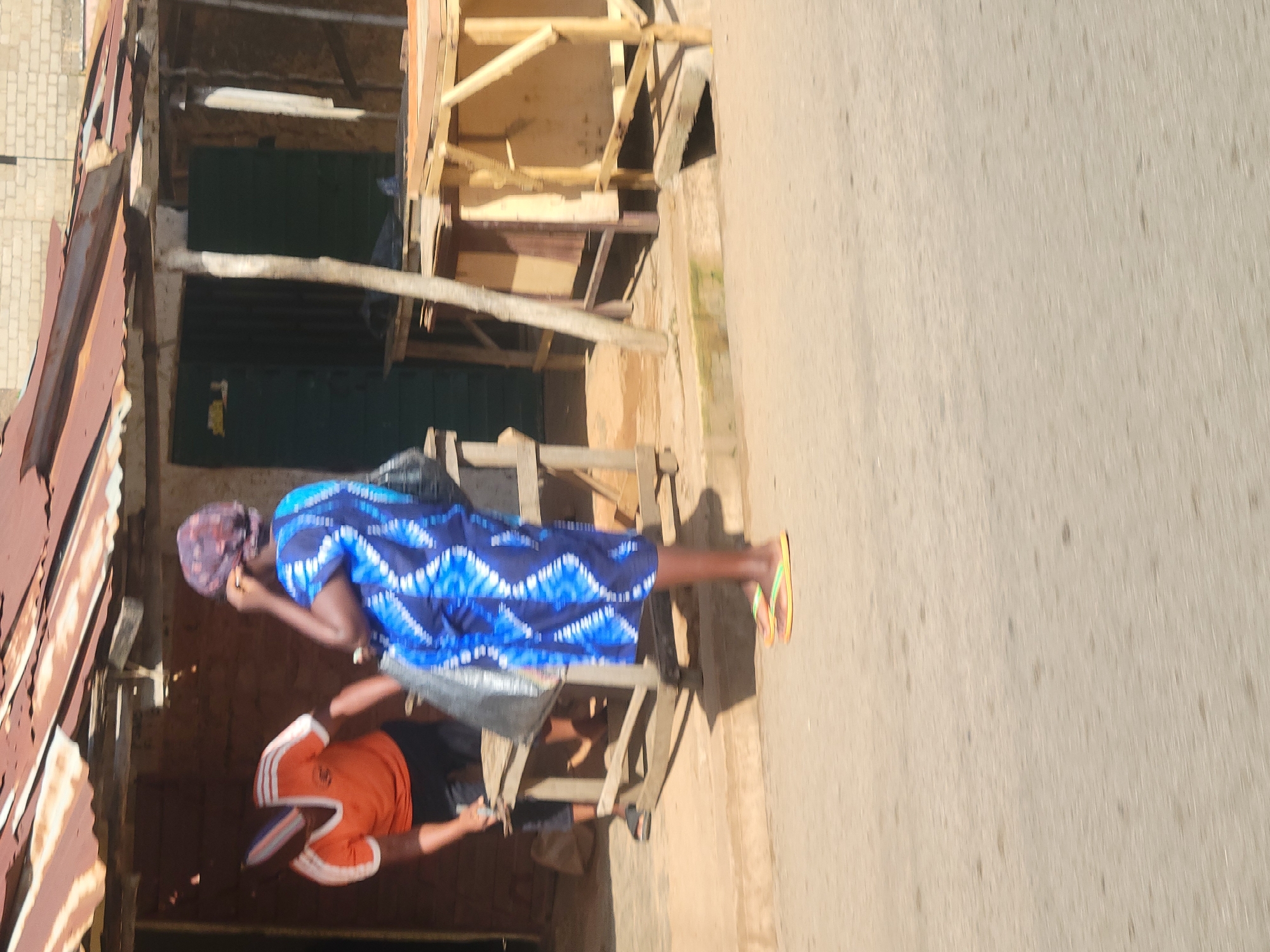 A medium shot captures an outdoor scene near a makeshift stall, likely in Jos, Nigeria, where two individuals are interacting. On the left, a person wearing an orange and white striped t-shirt and black pants is partially visible, leaning over a wooden structure, appearing to be engaged in an activity with a dark bag. To the right, a woman in a vibrant blue and white tie-dye dress and a headscarf stands with her back to the camera, holding a dark bag. Her feet are visible, wearing yellow and green flip-flops. The environment suggests a bustling street market, with a rusty, corrugated iron roof overhanging the stall. Wooden tables and makeshift structures are present, indicating a commercial setting. The ground is paved with concrete or asphalt, with some debris and a small drain visible. The overall lighting suggests daylight.