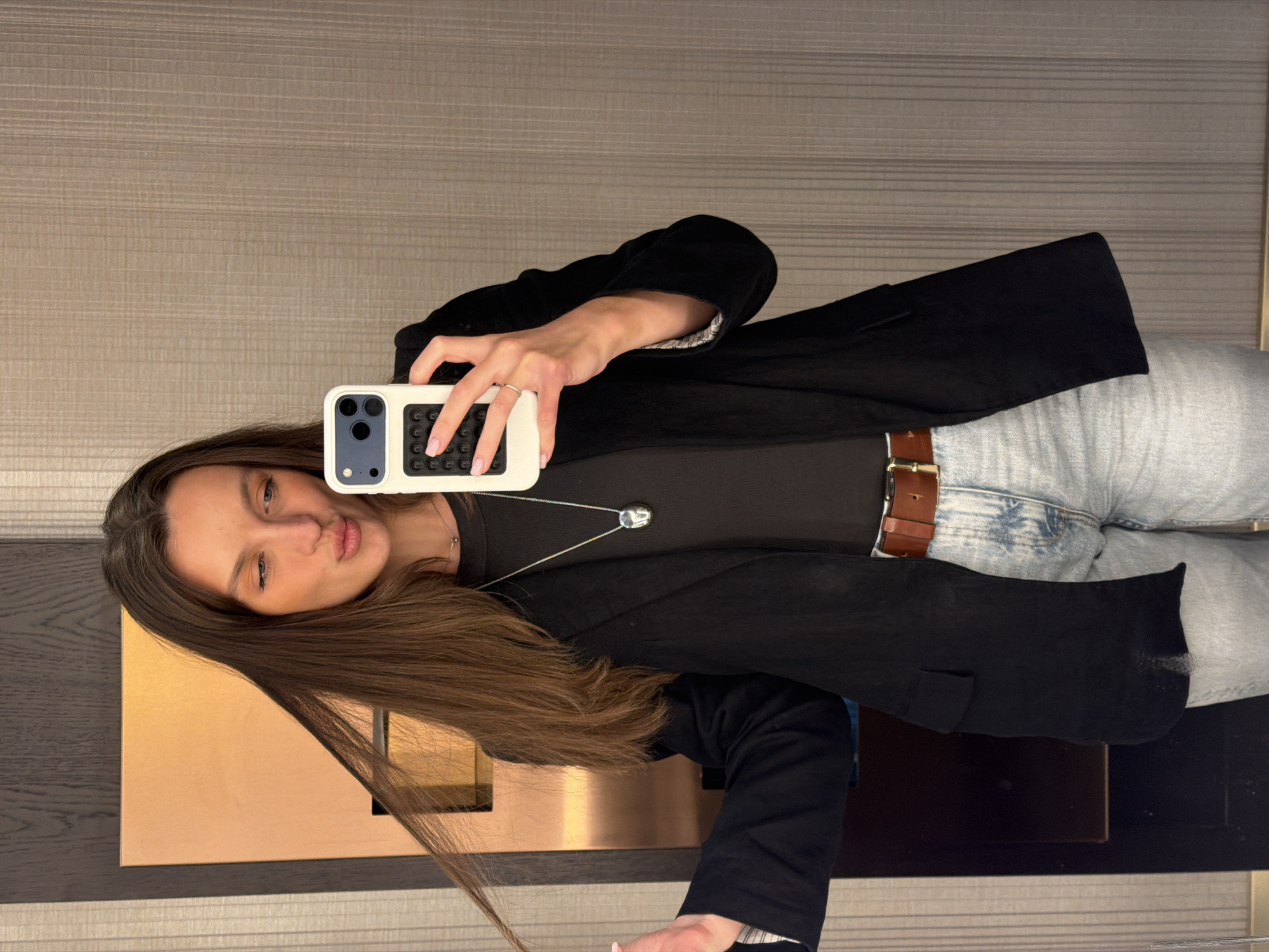 A woman is taking a selfie in a room with horizontally striped walls. She is wearing light wash jeans with a brown belt, a black t-shirt, and a black blazer. She is holding a white smartphone with a case that resembles a keypad. A silver necklace is visible around her neck. The lower portion of a wooden structure with a metallic front panel is visible in the bottom left corner of the image. It is not possible to determine the location in Budva, Montenegro, based on the image.