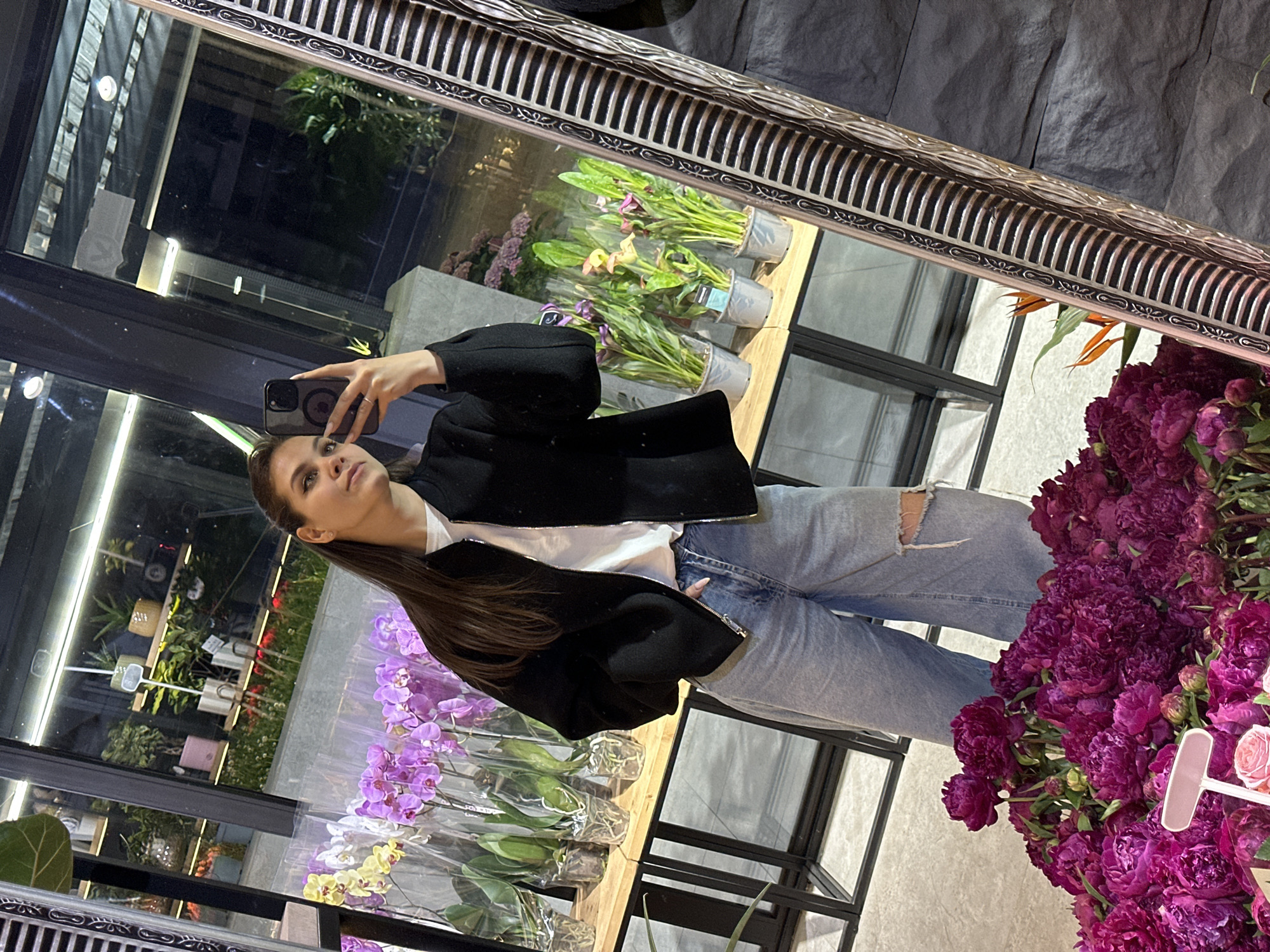 A young woman with long dark hair, wearing a black bomber jacket, a white t-shirt, and light-wash ripped jeans, stands in a flower shop in Chișinău, Moldova, taking a selfie in a large, ornate mirror. In front of her, a display of vibrant pink peonies is visible. The shop's interior is reflected in the mirror, showing shelves lined with potted plants, including orchids and calla lilies. The woman is holding her phone in her right hand, with her left hand resting on her hip. The mirror frame is intricately carved with a dark and metallic pattern.
