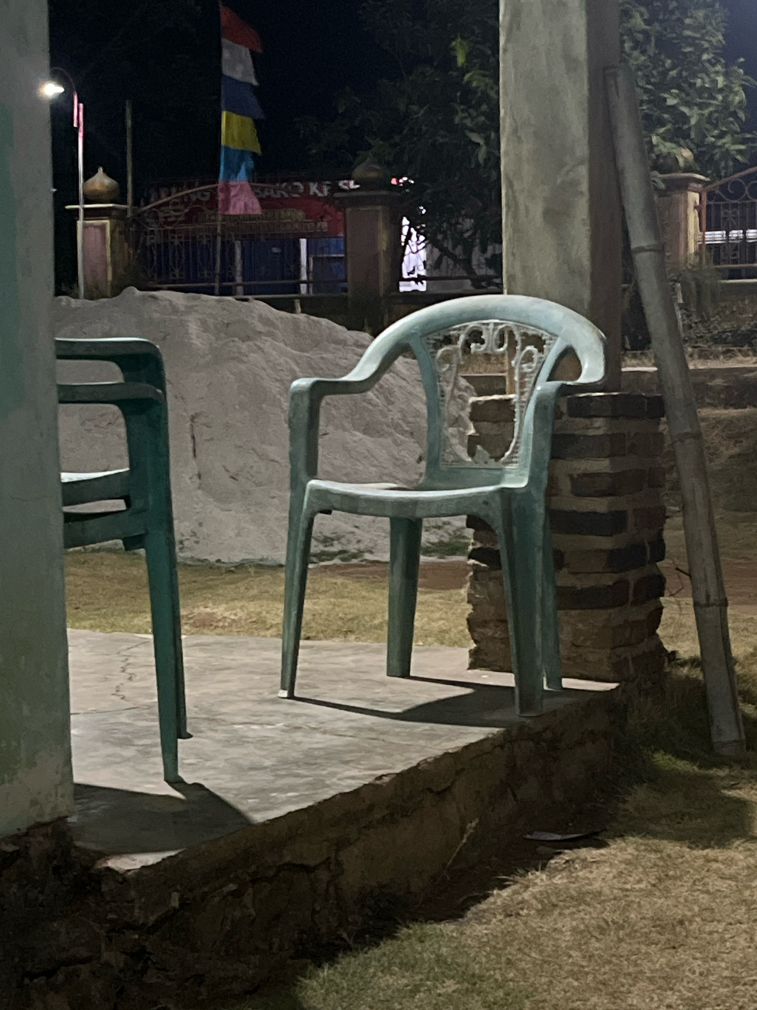 In the city of Kebumen, Indonesia, an outdoor scene is depicted at night. Two light blue plastic chairs are present; one is in the foreground to the left, mostly out of frame, and the other is centrally positioned, facing slightly to the right. The central chair is empty and rests on a raised concrete platform. Behind the chairs, a large pile of light-colored sand is visible, and beyond that, a fence with a gate and a sign with Indonesian text. A flag with horizontal red, white, yellow, blue, and pink stripes is displayed on a pole to the left of the gate. The area is lit by artificial light, casting shadows. The ground around the platform is covered with grass. A weathered brick pillar supports an overhead structure to the right of the central chair, with a long, light-colored pole leaning against it. The overall environment suggests an outdoor seating area, possibly at a construction site or a community gathering space.