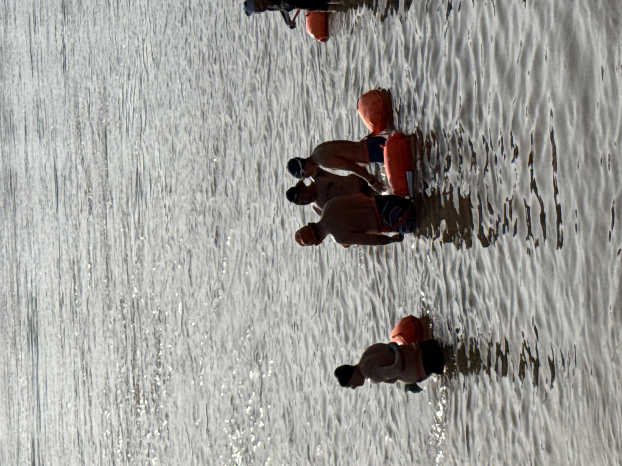 The image depicts a group of swimmers in the water near Jiangjin, China. Four shirtless men are wearing swim caps and appear to be treading water near bright orange buoys, while another man is on the far side of the frame. A fifth man wearing a hat and dark clothing is closer to the top of the frame and partially obscured. The water is textured with ripples and reflections of light.