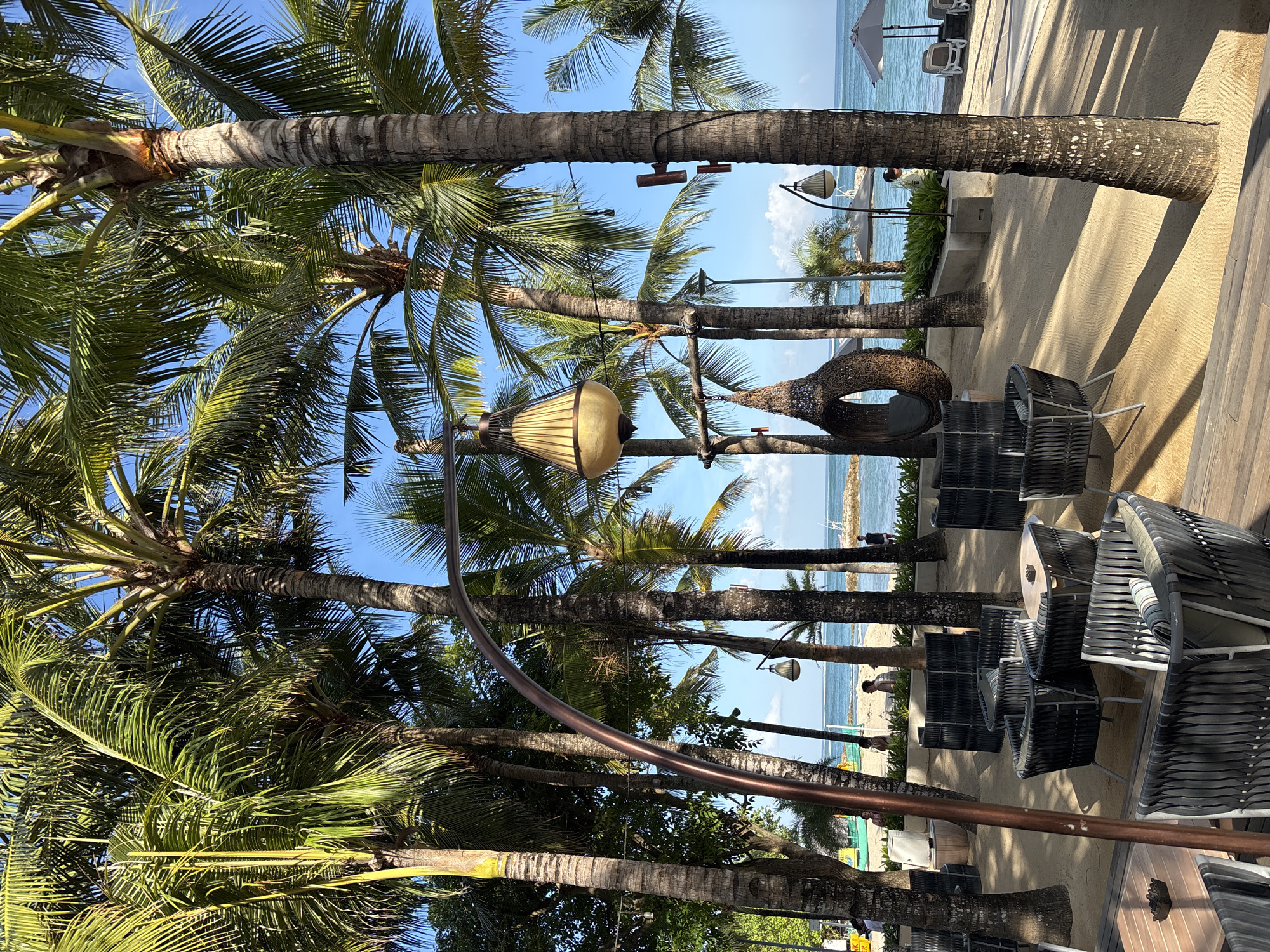 The image depicts a scenic outdoor seating area at a beachfront establishment in Kuta, Indonesia. The foreground features a wooden deck with dark gray, woven armchairs and tables arranged on a sandy surface. Beyond the seating area, a beach stretches towards the ocean, dotted with a few sun loungers and umbrellas. Tall palm trees with lush green fronds create a tropical ambiance, casting shadows on the sand. A woven egg-shaped swing hangs from a metal pole, adding a unique decorative element. In the background, the turquoise ocean meets a clear blue sky, with distant rocky outcrops visible. A few people are seen walking on the beach and near the water. The overall scene conveys a relaxed and inviting atmosphere, characteristic of a seaside resort or cafe in Kuta.