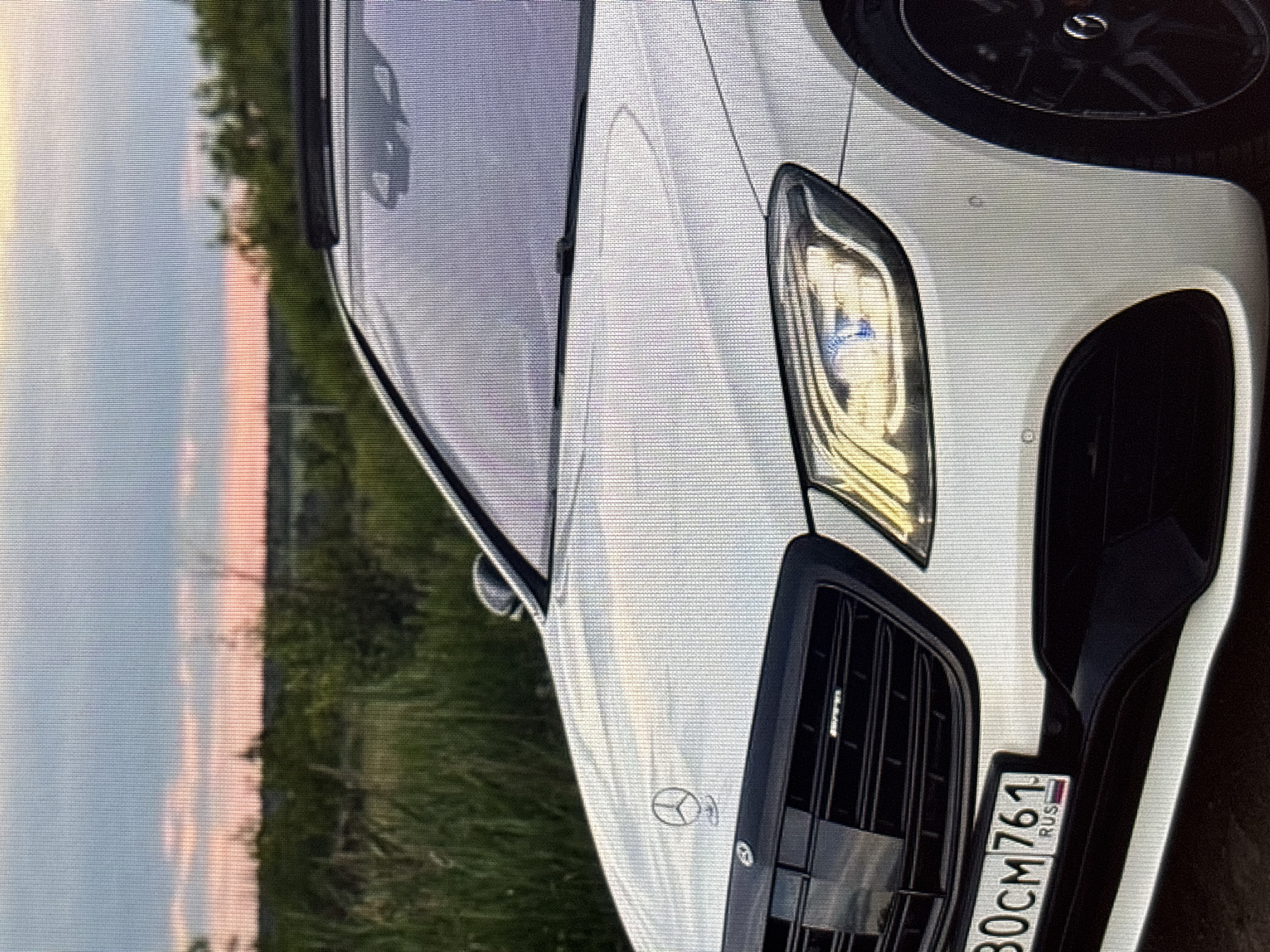 The image shows the front of a white Mercedes-Benz sedan parked outdoors. The car has a black grille with an AMG emblem and a license plate that reads "30CM 761 RUS." The sun is setting, casting a warm glow on the sky, and the car is surrounded by greenery. There are no people visible in the image. The image quality is low, with visible pixelation. No specific location in Moscow, Russia can be identified from the visual information provided.