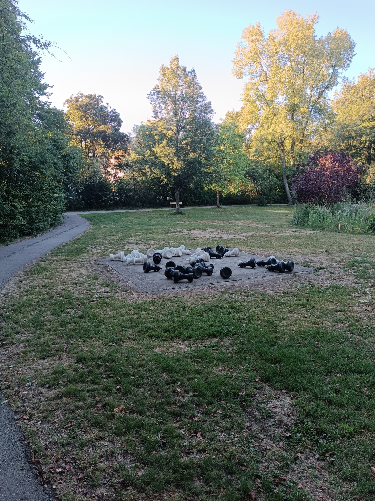 A set of oversized black and white chess pieces are scattered on a concrete slab in a grassy park. The pieces are lying on their sides, indicating a game that has been abandoned or disrupted. The park setting is characterized by mature trees with green and yellow foliage, a curved asphalt pathway, and a clear sky. The scene appears to be in the late afternoon or early evening, with soft sunlight illuminating the trees. This location is the Fünfseenpark in Fürstenfeldbruck, Germany.