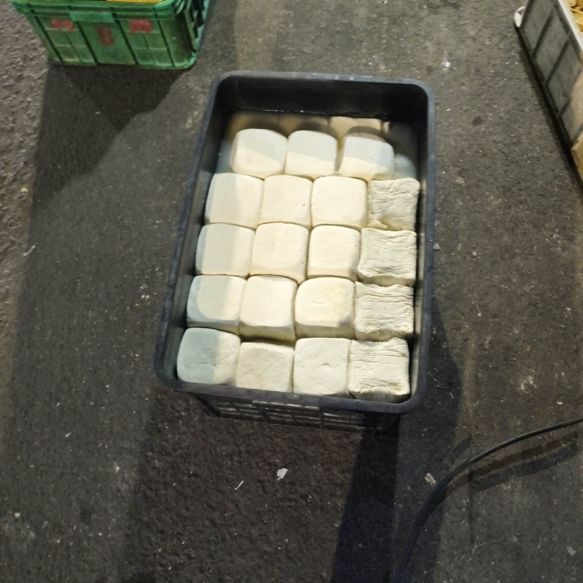 In South Jakarta, Indonesia, this image depicts a black plastic container filled with numerous squares of white tofu submerged in liquid, possibly water or brine. The container is placed on a dark, textured surface resembling asphalt or concrete. To the left, a green plastic crate is partially visible, containing yellow items. The lighting suggests an outdoor or partially covered environment, possibly a market stall, with shadows cast on the ground. The tofu is arranged in neat rows, with some pieces having a slightly folded or textured appearance, suggesting they might be packed or pressed in a specific way. The overall scene is static and appears to be a still life of food products.