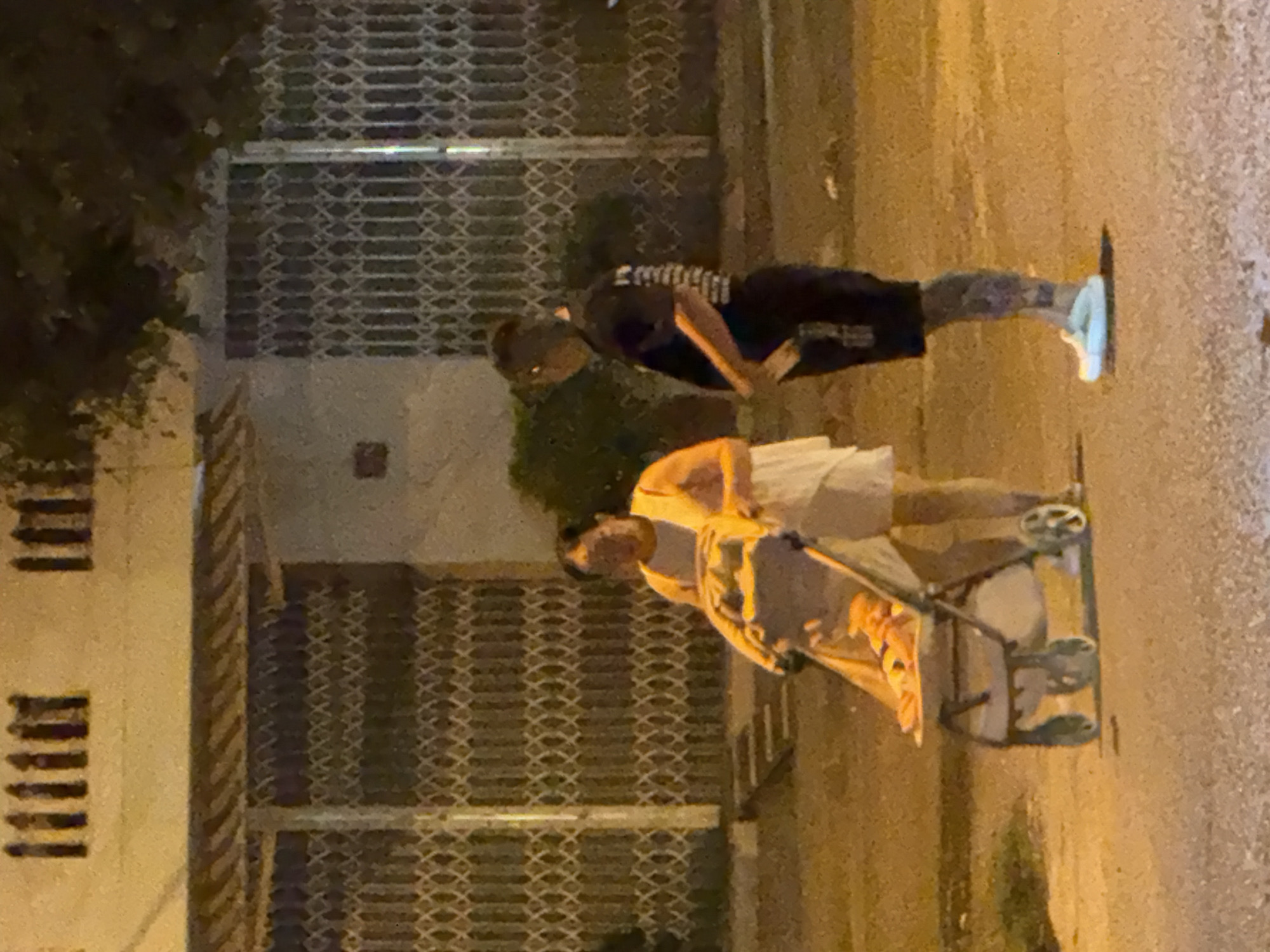 The image shows two people on a street at night. In the foreground, a person is pushing a stroller with a child inside. The child appears to be wearing a yellow top. In the background, another person is standing. They are wearing a dark t-shirt with white stripes on the sleeves and dark pants. The setting appears to be a street in Bac Nha Trang Ward, Vietnam, with a building and a metal gate visible. The street is paved with asphalt and the lighting suggests it is nighttime, possibly illuminated by streetlights. The overall scene is dimly lit, with a yellowish cast from the artificial light sources. The atmosphere seems ordinary, suggesting a casual moment of people out and about in their neighborhood. No discernible text is visible in the image.