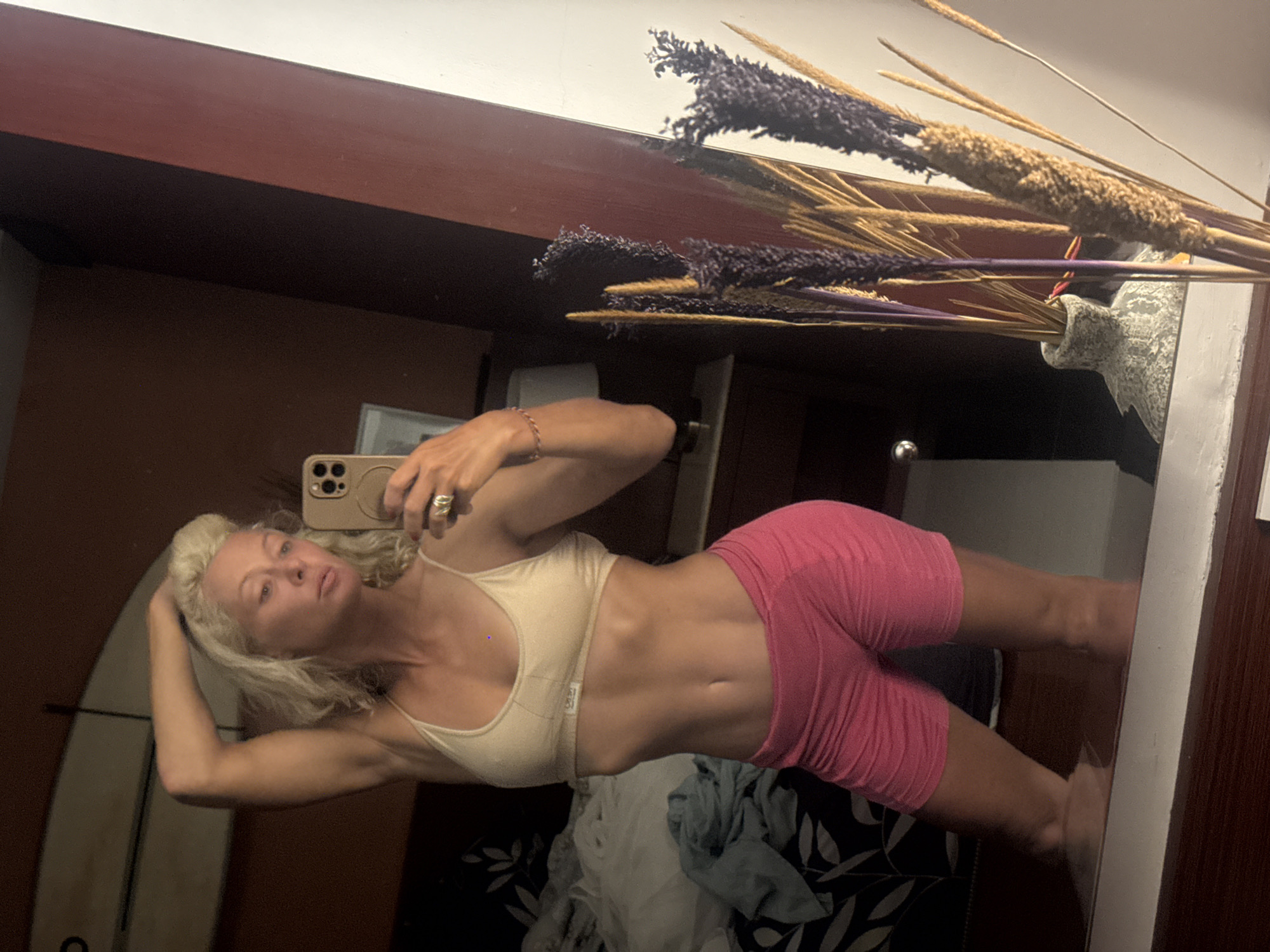 A woman is taking a selfie in a mirror. She is wearing a beige sports bra and pink shorts. Her blonde hair is loose. In the background, there is a surfboard, some clothing, and a vase with dried flowers. The location is Unknown, Unknown.