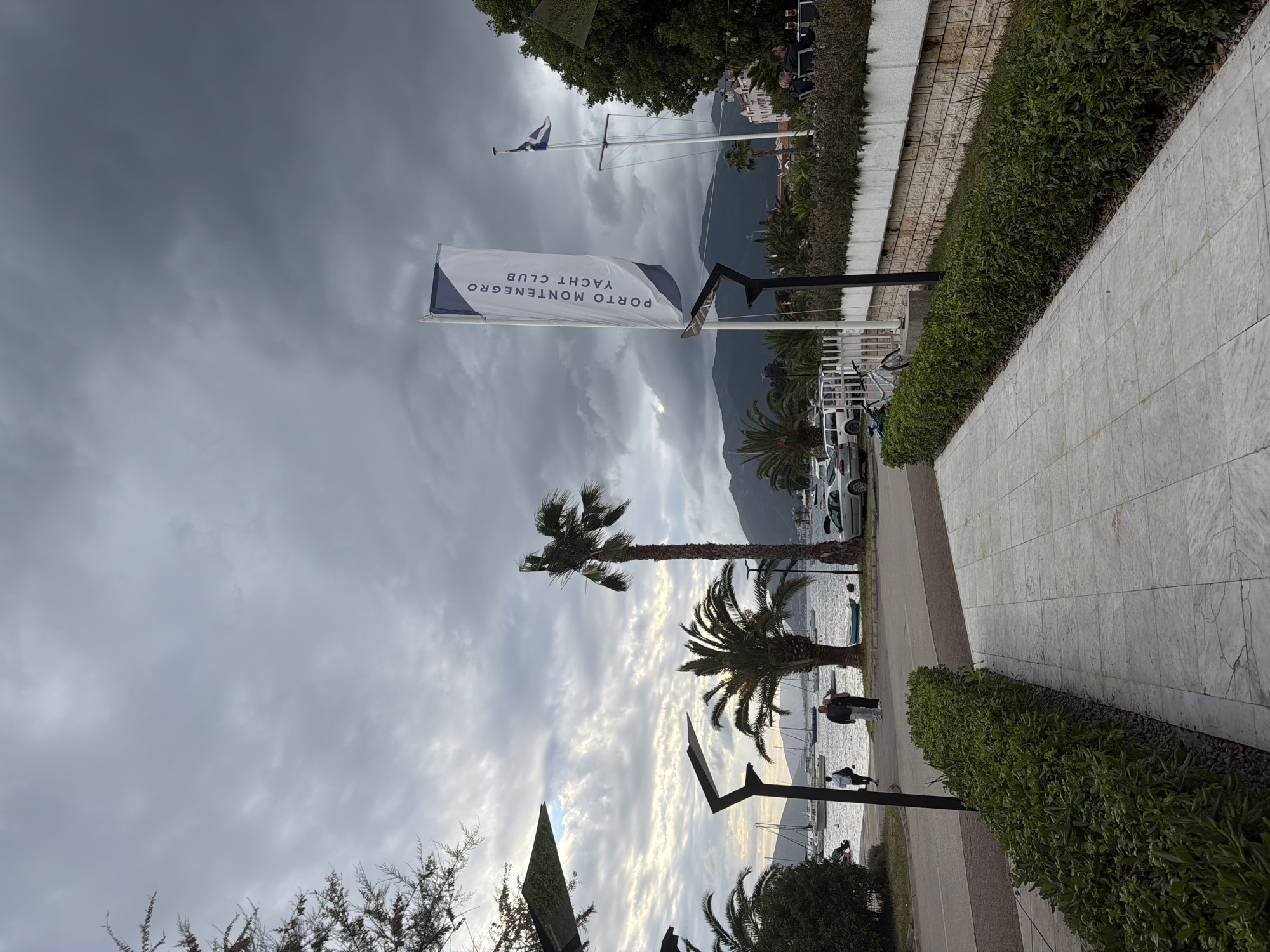 The image captures a view of Porto Montenegro Yacht Club in Tivat, Montenegro. The sky is overcast with dark clouds. A flag bearing the words "PORTO MONTENEGRO YACHT CLUB" is visible. Palm trees line the waterfront, and in the distance, mountains are discernible. Several people are walking along a paved path adjacent to the water. A row of cars and a building can be seen further down the road. A stone walkway is on the right side of the photo. A hedge borders the walkway.