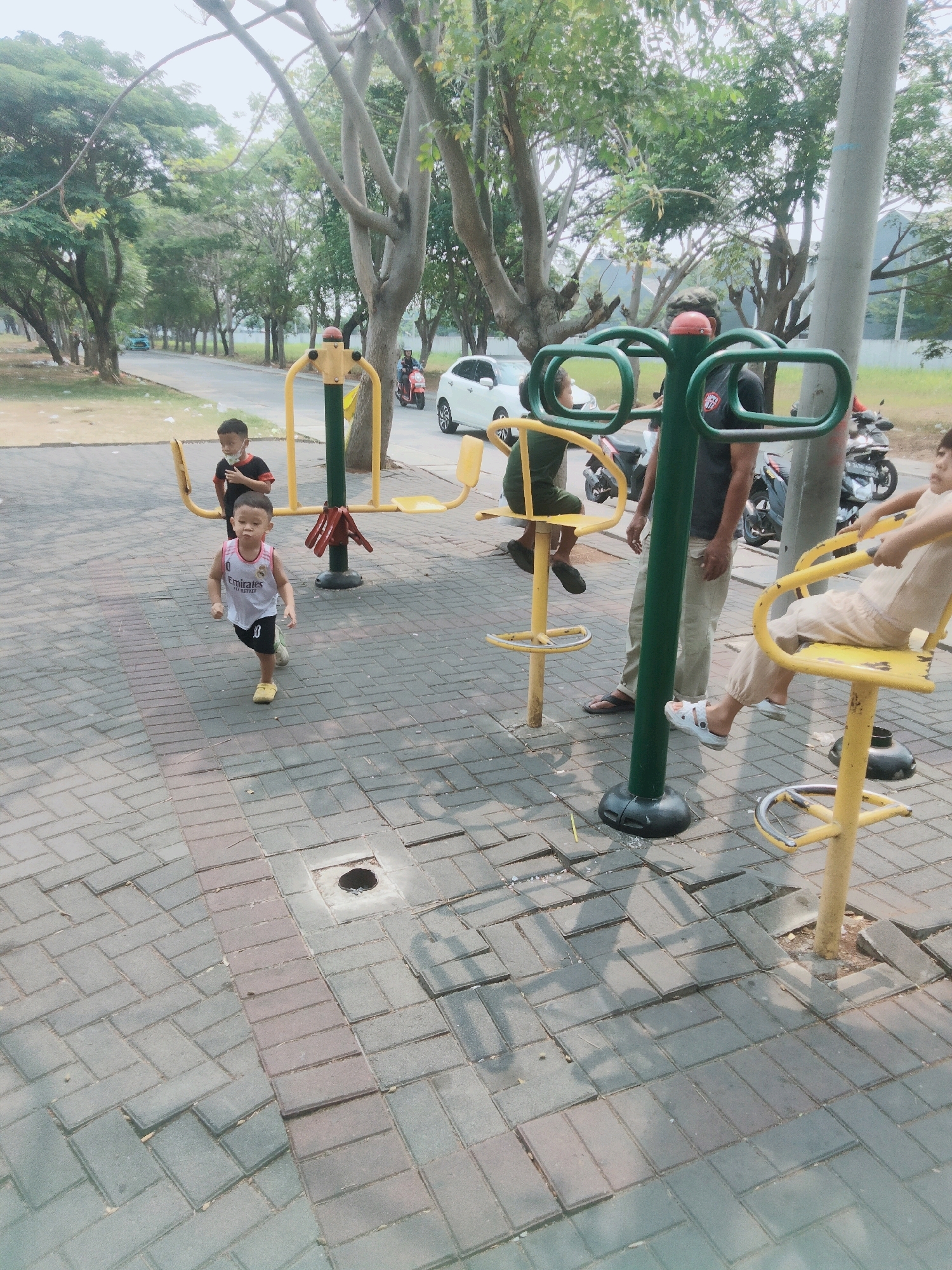 In a public park in Tangerang, Indonesia, a group of people are using outdoor exercise equipment. A young boy in a white jersey with black trim is running on the paved ground in the foreground. Behind him, another boy in a black and red shirt stands near a yellow and green exercise machine. Several adults are also present, some seated on exercise equipment. Motorcycles are parked in the background, and a white car is visible on the road. The environment is a park with trees and a paved walkway, indicating an urban setting.