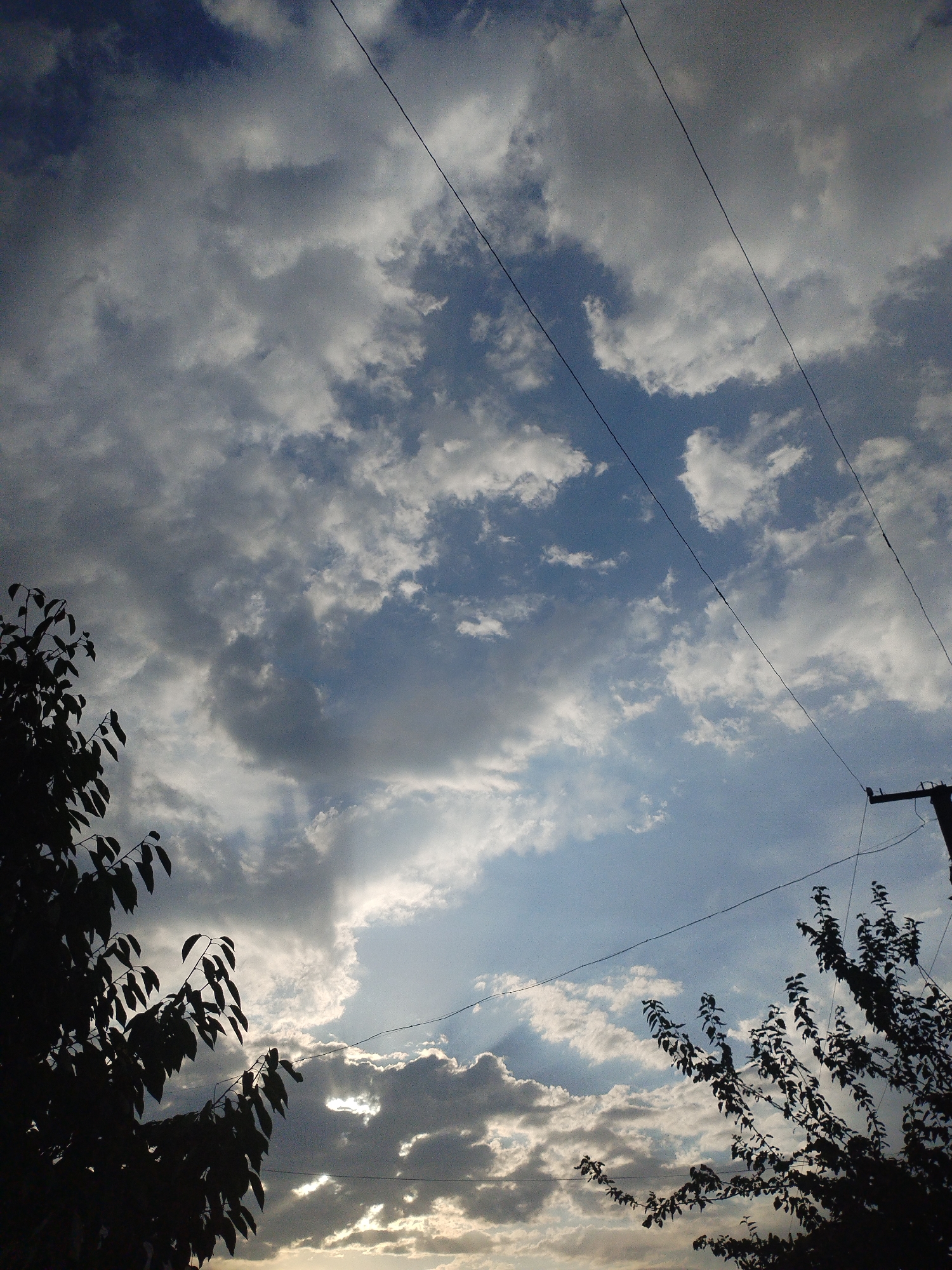 The image depicts a cloudy sky with the sun partially visible behind the clouds, suggesting it is either early morning or late afternoon. Silhouetted against the sky are the branches of trees on the left side of the frame and on the right side. Two power lines stretch diagonally across the image. On the far right, a utility pole is visible. The scene appears to be an outdoor setting, possibly in a residential area or a park, given the presence of trees and utility infrastructure. The location is identified as Kyzyl-Kyya, Kyrgyzstan. There are no people or animals visible in the image, nor any specific actions or interactions taking place. The environment is characterized by the natural elements of sky and vegetation, along with man-made structures like power lines and a pole.