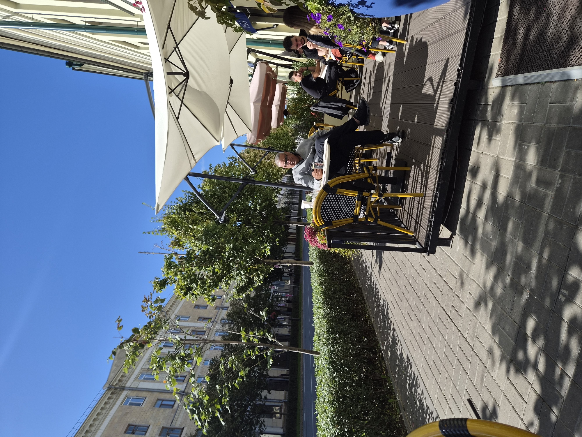 A group of people are seated at tables outside a cafe on a sunny day. The cafe is located on a street in Kazan, Russia, as indicated by the architectural style of the buildings and the presence of birch trees, which are common in the region. The people are casually dressed and appear to be enjoying their meals or drinks. One man in the foreground is wearing a gray sweatshirt and black pants, and he is holding a glass of water. A woman in the background is wearing a blue dress, and she has long, dark hair. The cafe has white umbrellas to provide shade for the customers. The street is lined with trees and a hedge, and there is a sidewalk with a brick pattern. The sky is clear and blue.