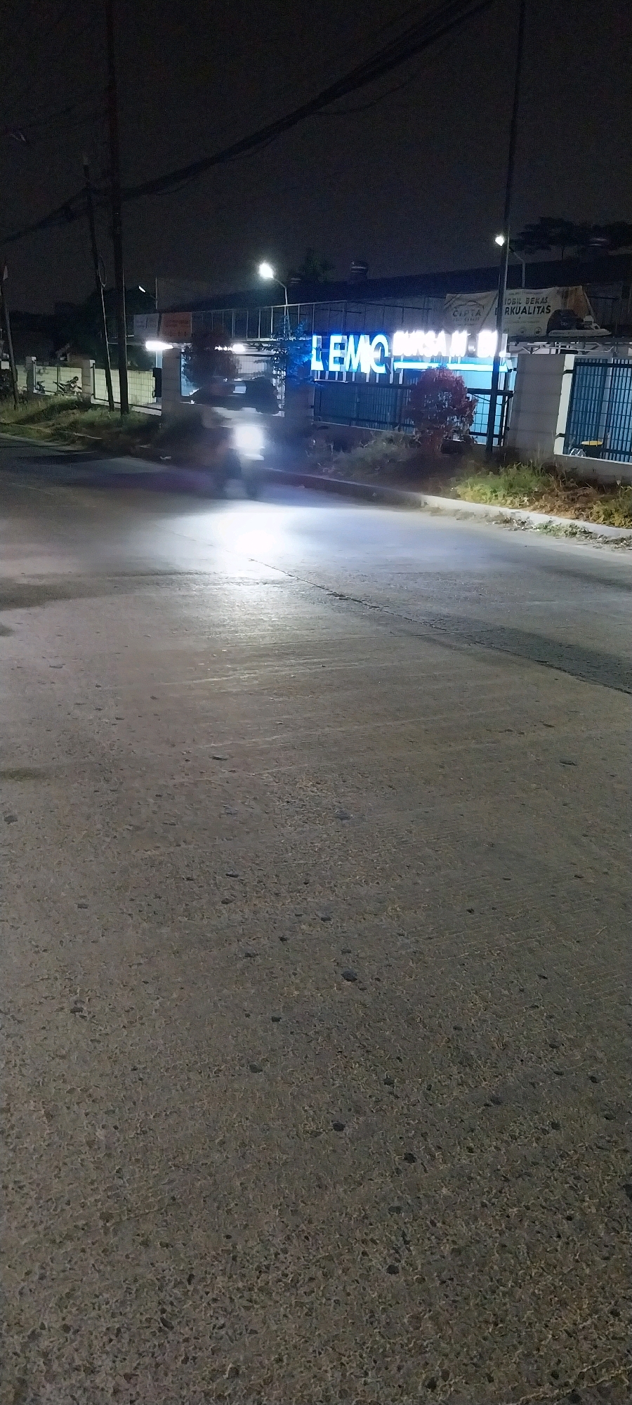 The image depicts an outdoor scene at night, likely in Kelapa Dua, Indonesia, based on the included text. A paved road occupies the foreground and midground, with a motorcycle producing bright headlights moving away from the viewer towards a building in the background. The building features a prominent illuminated sign that reads "LEMO" in large blue letters, with additional text partially visible and appearing to say "BURSA MOBIL BEKAS BERKUALITAS" in smaller lettering above and to the right of the "LEMO" sign, which translates to "Quality Used Car Exchange." The building itself is modern with metal fencing and a glass facade. There are streetlights and overhead power lines visible in the darker sky. Vegetation is present along the side of the road and in front of the building. The overall impression is of a commercial area illuminated for nighttime operation.