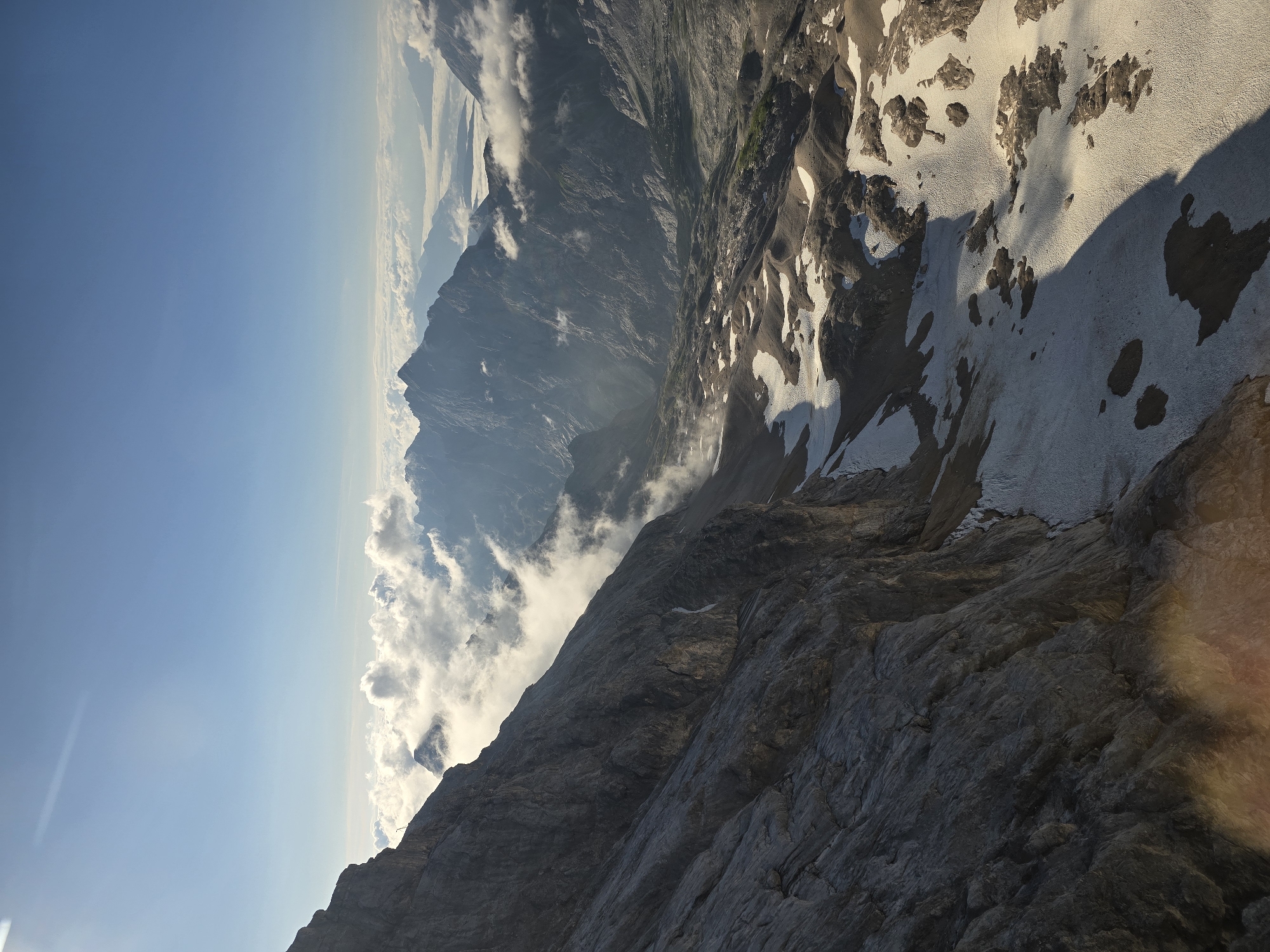 The image depicts a mountainous landscape, likely in the Alps. In the foreground, a steep, rocky mountain face dominates the left side of the frame, with patches of snow scattered across its surface. To the right, a vast valley stretches out, filled with clouds that obscure the lower terrain. Beyond the valley, a series of jagged mountain peaks rise, their silhouettes softened by the distance and the atmospheric haze. The sky above is a clear blue, with the sun casting a bright light, creating lens flare in the lower left corner and illuminating the clouds. This scene is likely a high-altitude vista, possibly taken from a summit or a cable car, offering a panoramic view of the alpine region. The location is identified as the Zugspitze massif, near Garmisch-Partenkirchen, Germany, known for its dramatic mountain scenery and glacial remnants. There are no people or animals visible in the image, only the raw, natural elements of the mountain environment.