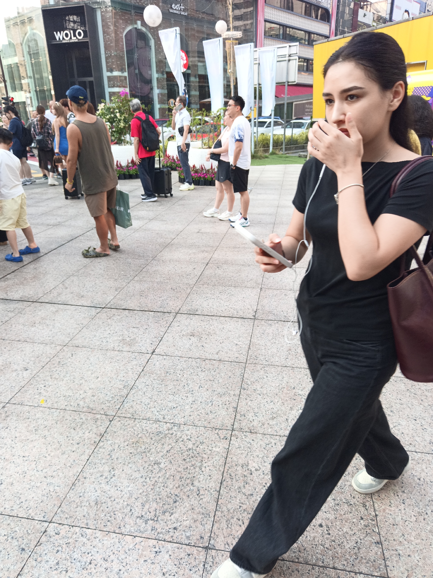 A woman is walking on a tiled sidewalk in a city. She is wearing a black t-shirt, black pants, and white sneakers. She has earphones in her ears and is holding a smartphone in her right hand. A brown purse is slung over her left shoulder. In the background, several other people are walking, some with luggage. Buildings line the street, one of which has a large "WOLO" sign. The environment appears to be a busy commercial district, likely in Kuala Lumpur, Malaysia, given the signage.