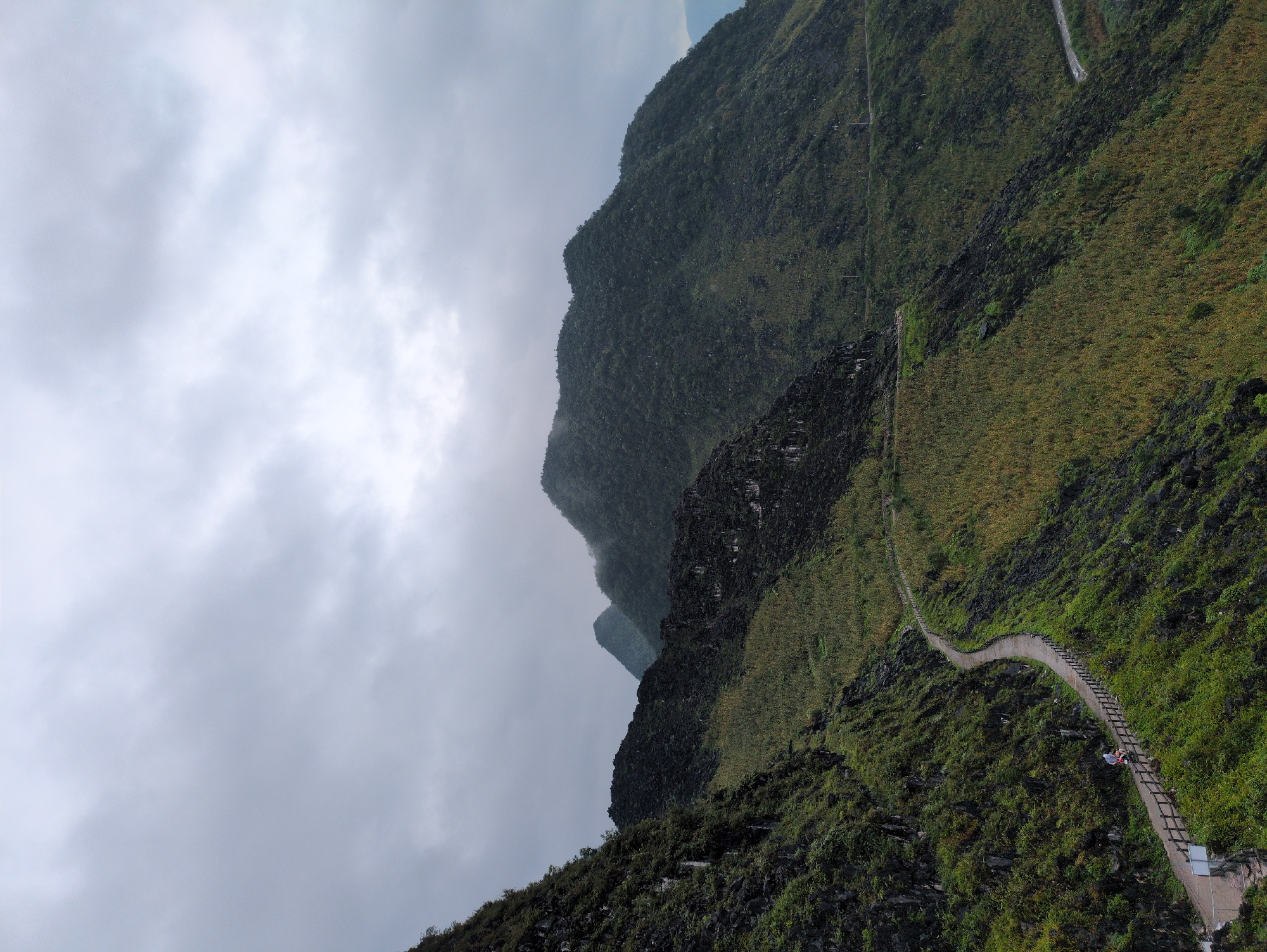 The image shows a winding road on a mountain slope in Đồng Văn Commune, Vietnam. The road is paved and bordered by a wooden fence on the left side. A person is riding a motorcycle on the road, heading away from the viewer. The mountain is covered in lush green vegetation, with some rocky outcrops visible. In the background, several other mountains are visible, shrouded in mist. The sky is overcast, with heavy clouds. The scene depicts a rural, mountainous landscape with a focus on the winding road and the surrounding natural environment.