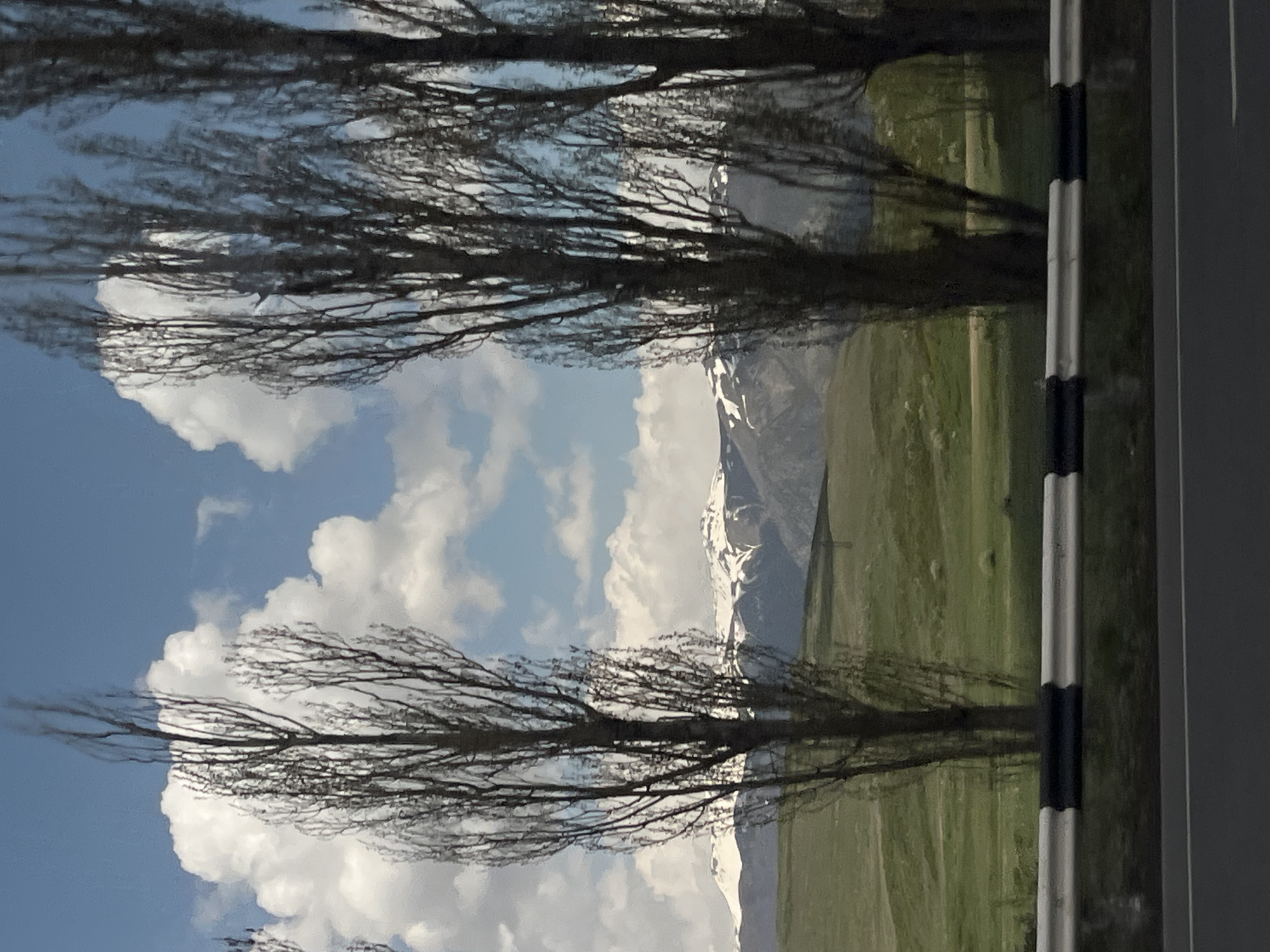 A landscape photograph captures a picturesque view in Charentsavan, Armenia. The scene features a mountain range partially covered in snow, which is visible through gaps in the branches of trees. The trees, with their bare branches, extend across the frame against a backdrop of a blue sky dotted with white clouds. To the right, a roadside barrier is visible, marked with black and white stripes. A field of green stretches towards the mountains. The composition suggests the photograph was taken from a vehicle.