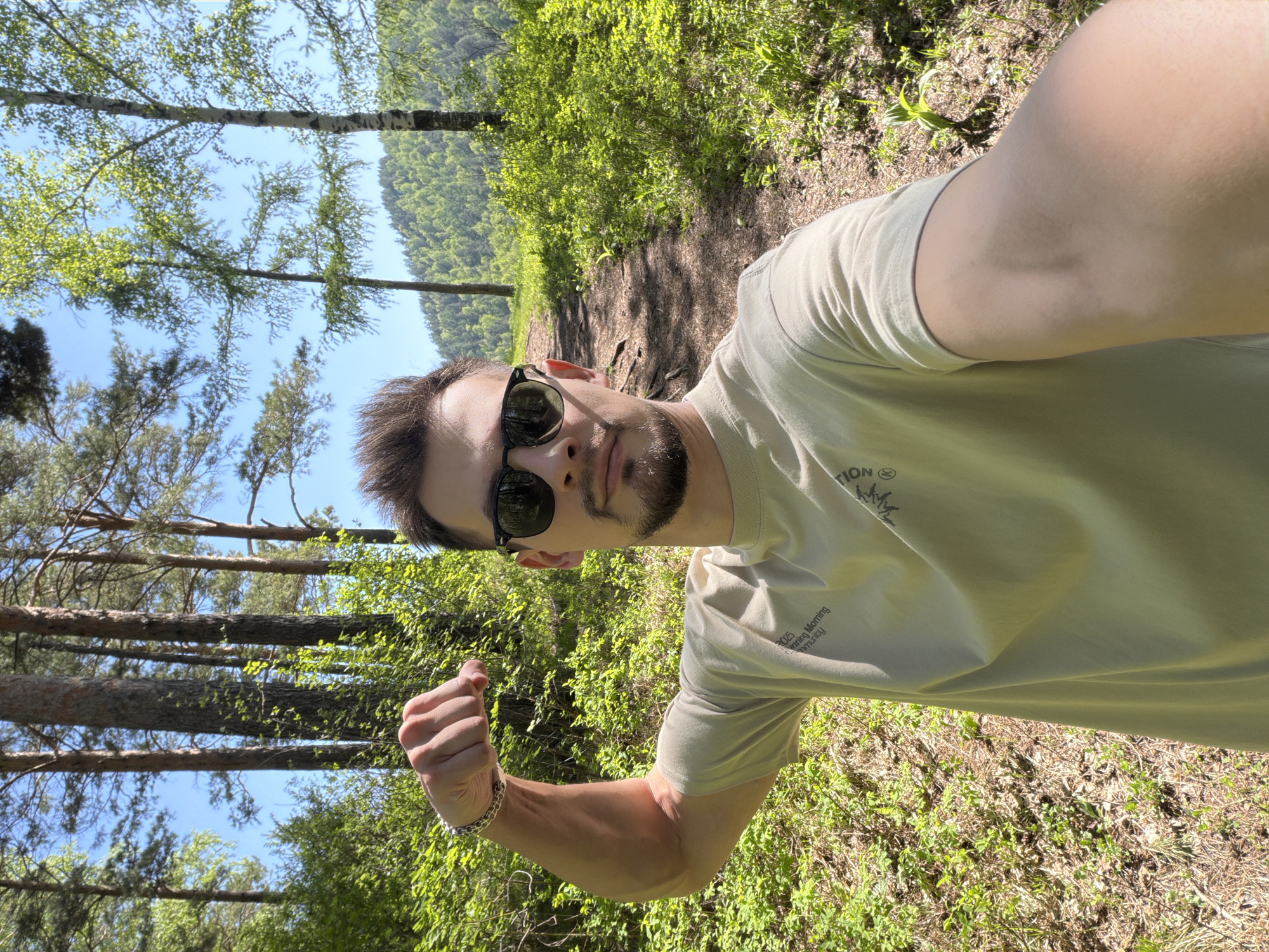 This media file is a bright, sunlit outdoor selfie, rotated 90 degrees clockwise. The main subject is a young, fair-skinned man with short dark hair, a beard, and sunglasses, looking directly at the camera. He is wearing a light-colored t-shirt, which has dark text visible on the chest: "TION" and below it, "2023 Morning Community". The man's left arm is flexed, forming a fist, showcasing his bicep, and he wears a silver chain bracelet on his wrist.
The setting is a vibrant natural environment, indicative of a forest or wooded trail in Krasnoyarsk, Russia. The background is filled with lush green foliage, including tall trees with light-colored bark (resembling birch) and darker evergreen trees, stretching upwards towards a clear, bright blue sky. A dirt path or natural ground covered with green plants and some dried leaves is visible, suggesting a hiking or walking area. The bright lighting and clear sky indicate daytime, likely a sunny morning or afternoon. The image conveys an active, outdoor moment.