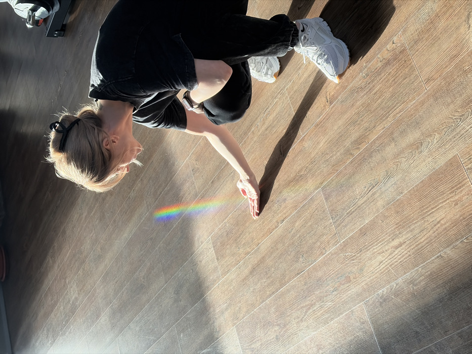 An individual, appearing female, with light-colored hair styled in a bun with a black claw clip, is captured in a crouched position, extending her right hand with the palm facing upwards. She wears a black short-sleeved t-shirt, dark-colored sweatpants, and white athletic shoes with light brown soles. A dark-colored smartwatch is visible on her left wrist. The scene takes place indoors on a floor covered with light-to-medium brown wood-look plank flooring. A distinct, vertical rainbow spectrum of light, originating from an unidentifiable source outside the frame, is projected onto the floor, directly intersecting with her outstretched hand. Strong, directional natural light illuminates the scene from the left, casting shadows. In the upper right background, a portion of dark-colored exercise equipment is partially visible. The specific location within Chaikovsky, Russia, is not identifiable from the image.